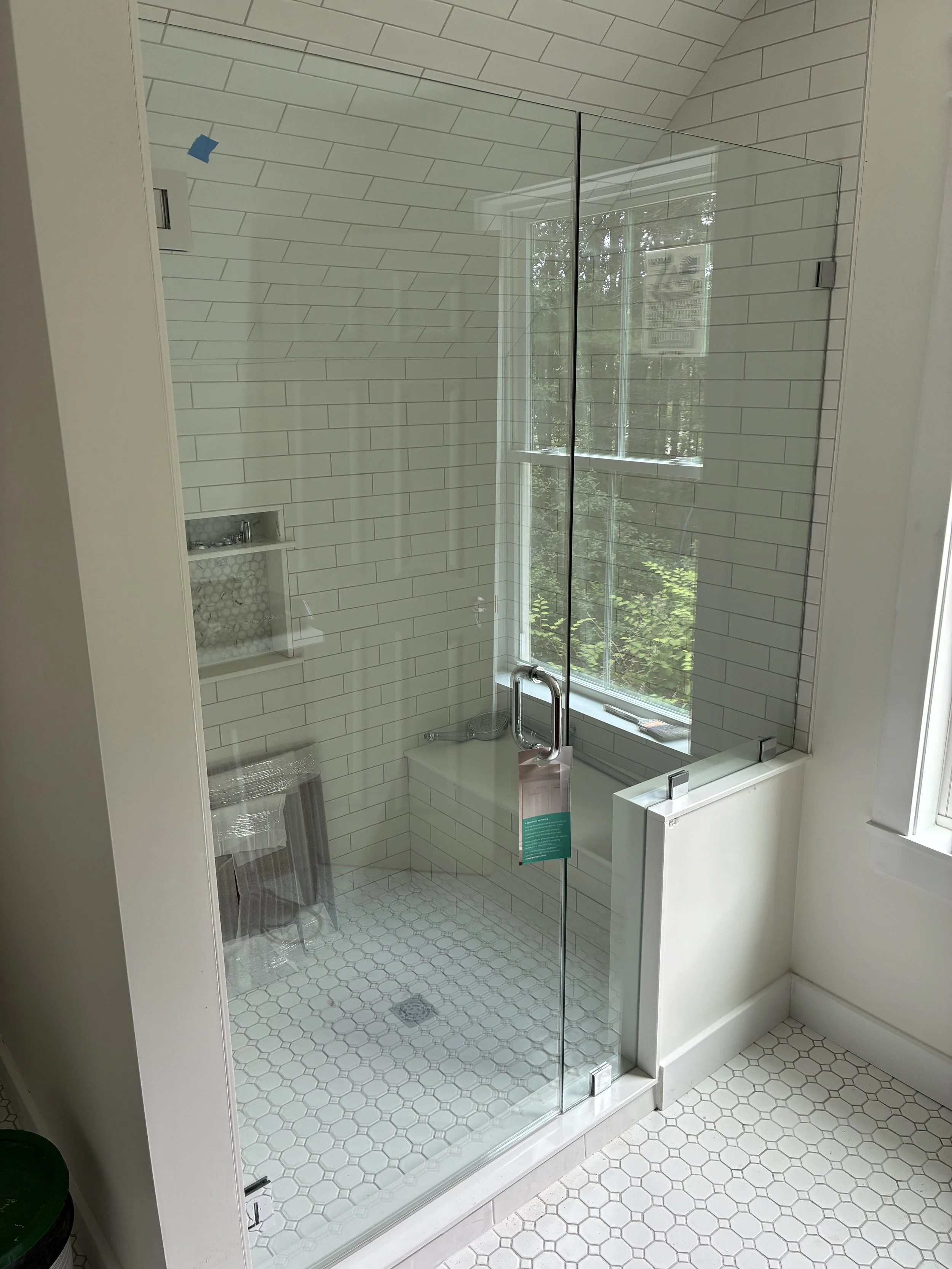 A glass enclosed shower with white subway tile walls, a small window, a built-in shelf with hexagonal tile backing, and a chrome shower handle. Outside the shower are white hexagonal floor tiles and a partial view of a window with greenery outside.