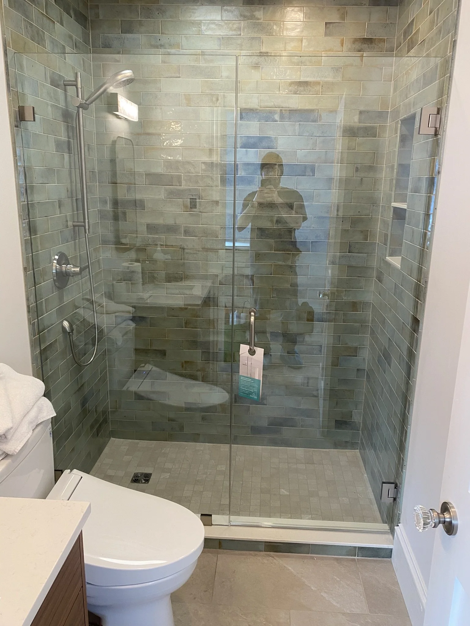 Bathroom with a glass-enclosed shower, a white toilet, and beige tiled flooring and walls. The shower has greenish tiles and a sliding glass door, with a person’s reflection visible taking a photo.