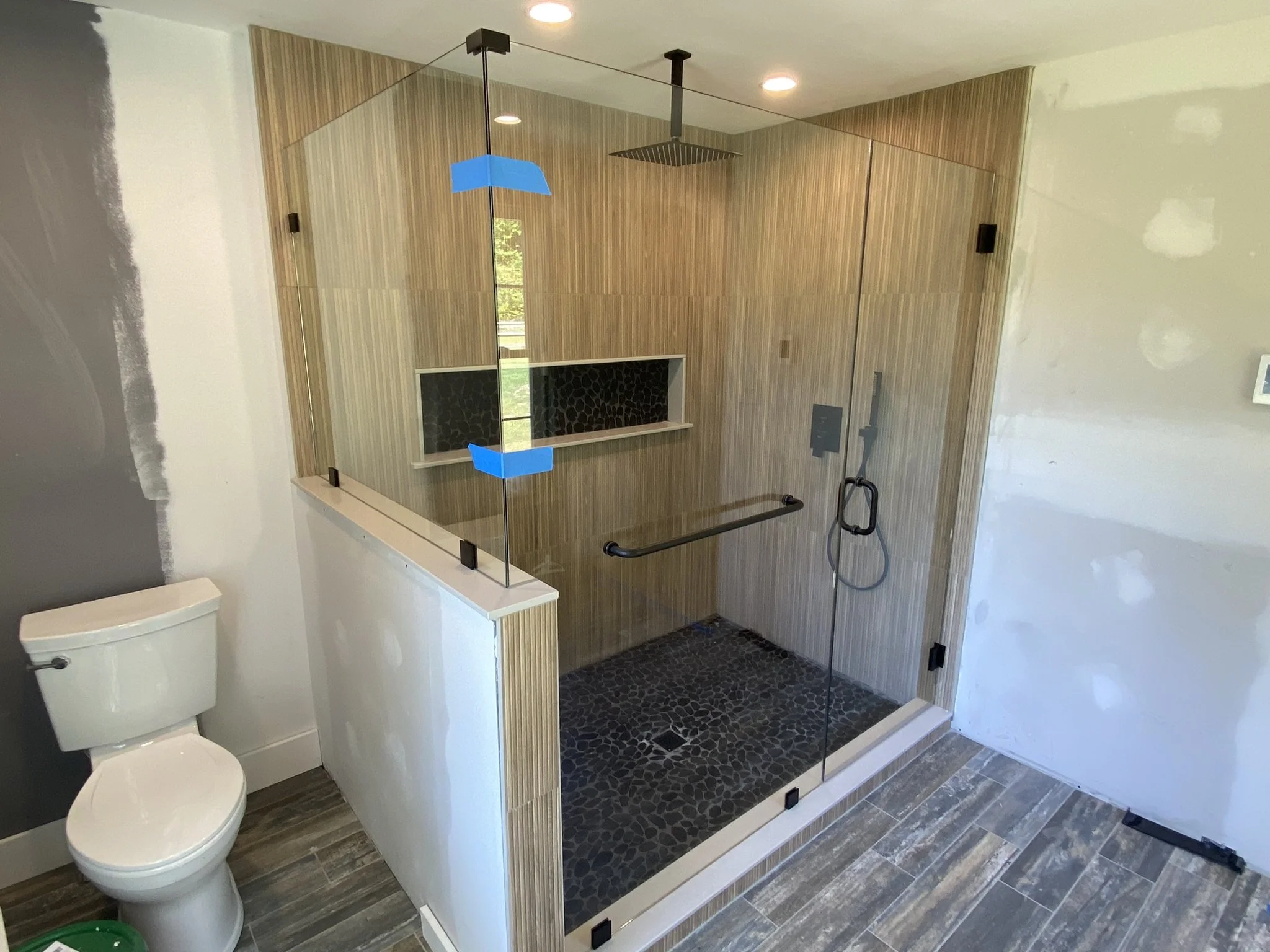 A bathroom with a glass-enclosed shower area, a white toilet, and wood paneling on the shower walls. The shower has black pebble-style flooring, a black safety grab bar, and a black and gray patterned built-in niche. The ceiling has recessed lights, 