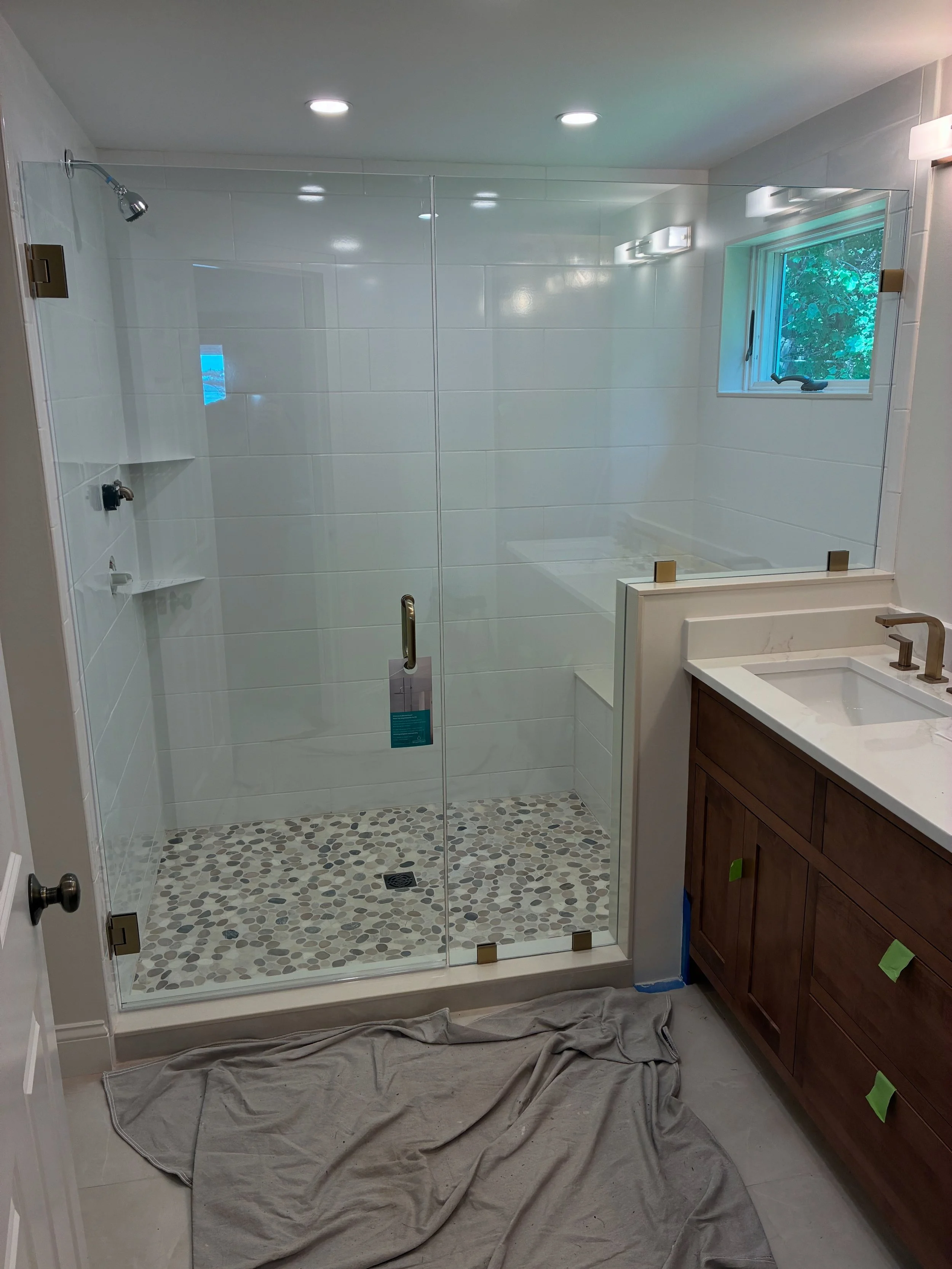 A modern bathroom with a glass-enclosed shower that has pebble flooring, a small window, white tile walls, and a wooden vanity with a white countertop and bronze fixtures.