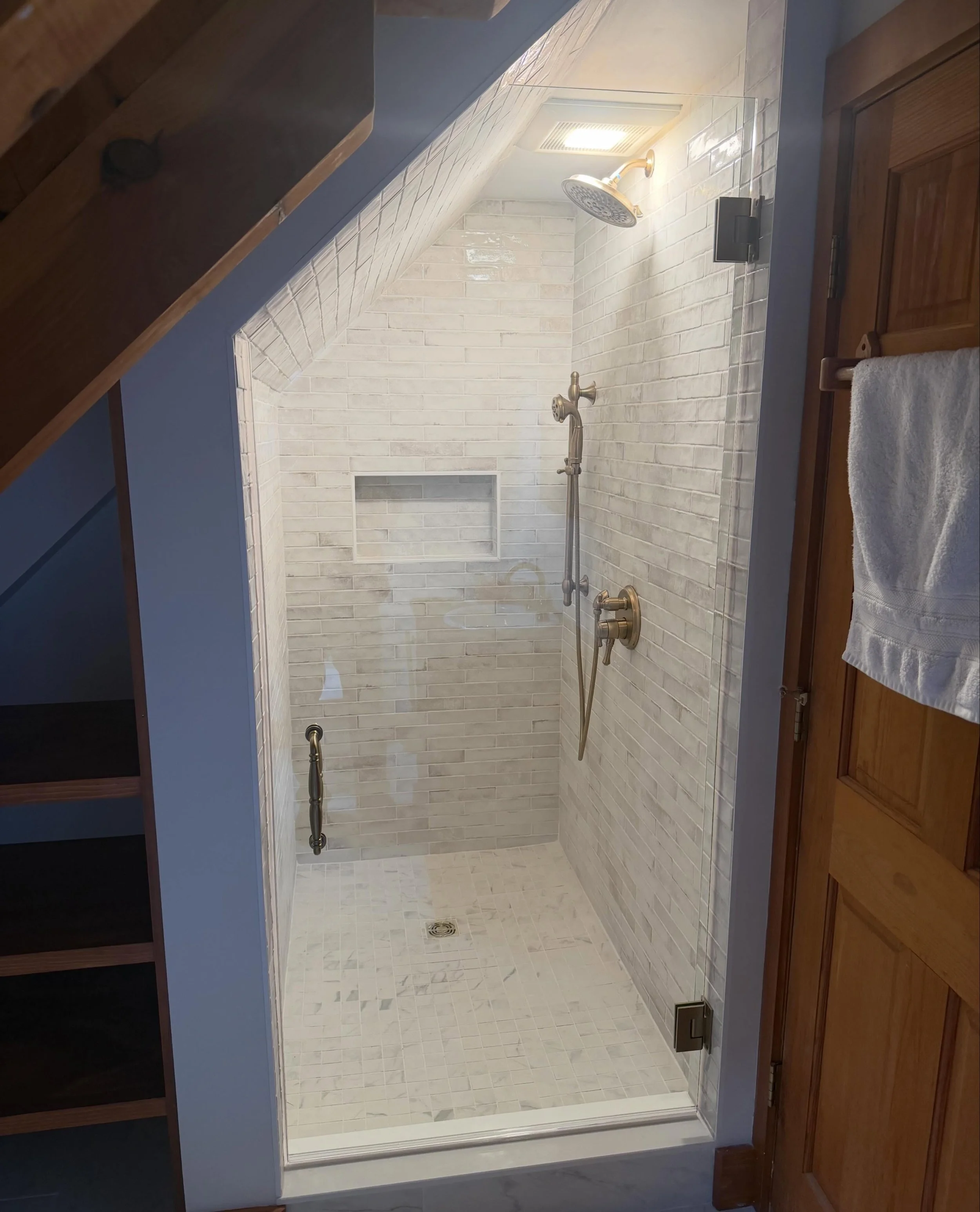 Frameless glass shower door and panel cut to a sloped ceiling under a staircase, with brass fixtures, white marble-look tile, a niche and a mosaic floor, in a Buxton, Maine bathroom