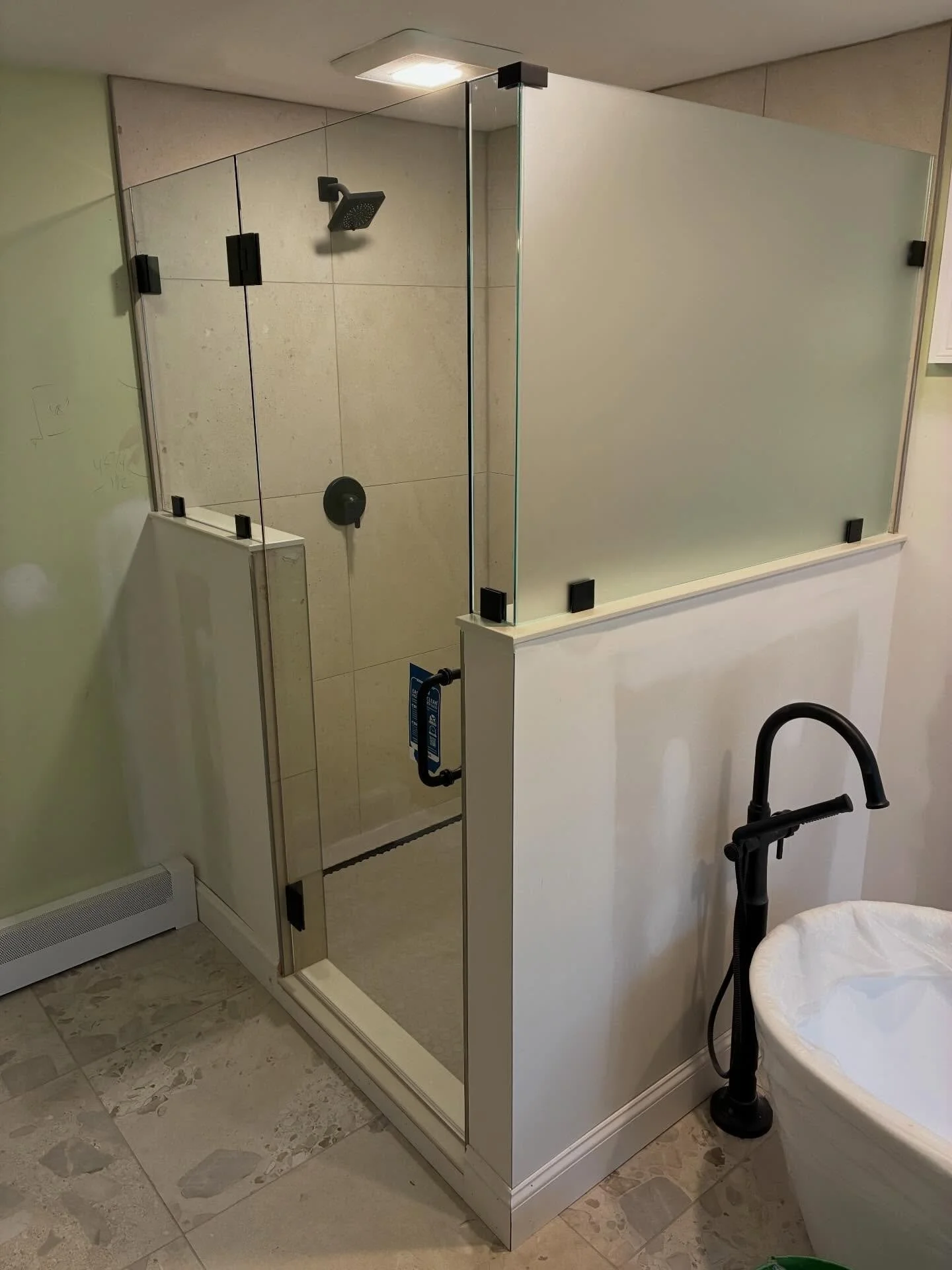 Frameless glass shower enclosure with a clear hinged door and front panel and a frosted privacy glass panel on a white knee wall, matte black clips, beside a freestanding tub in a Falmouth Foreside, Maine bathroom