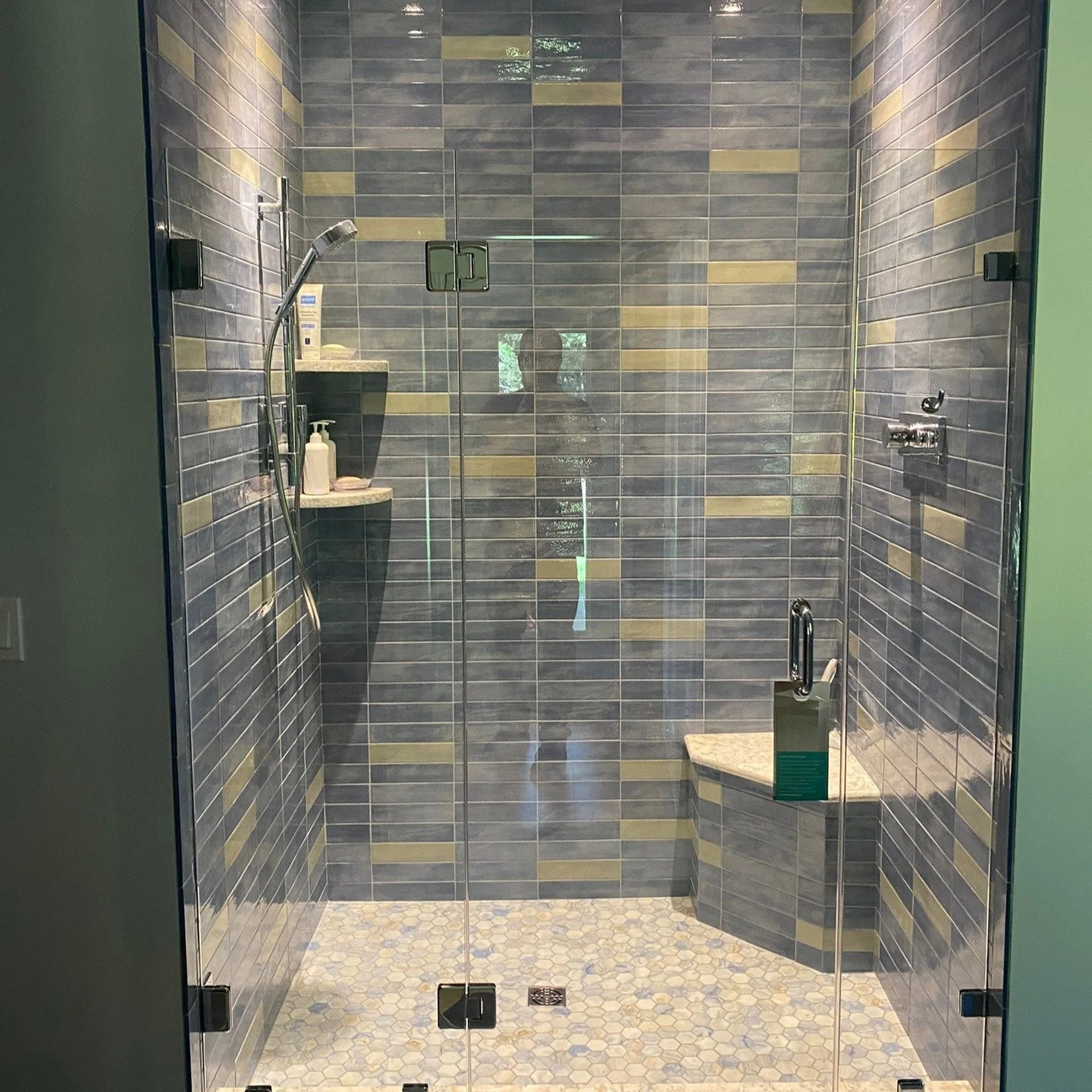 Frameless glass shower door and panels with matte black hinges and clips, blue and gold tile, a corner bench and a hexagon mosaic floor, in a Yarmouth, Maine bathroom