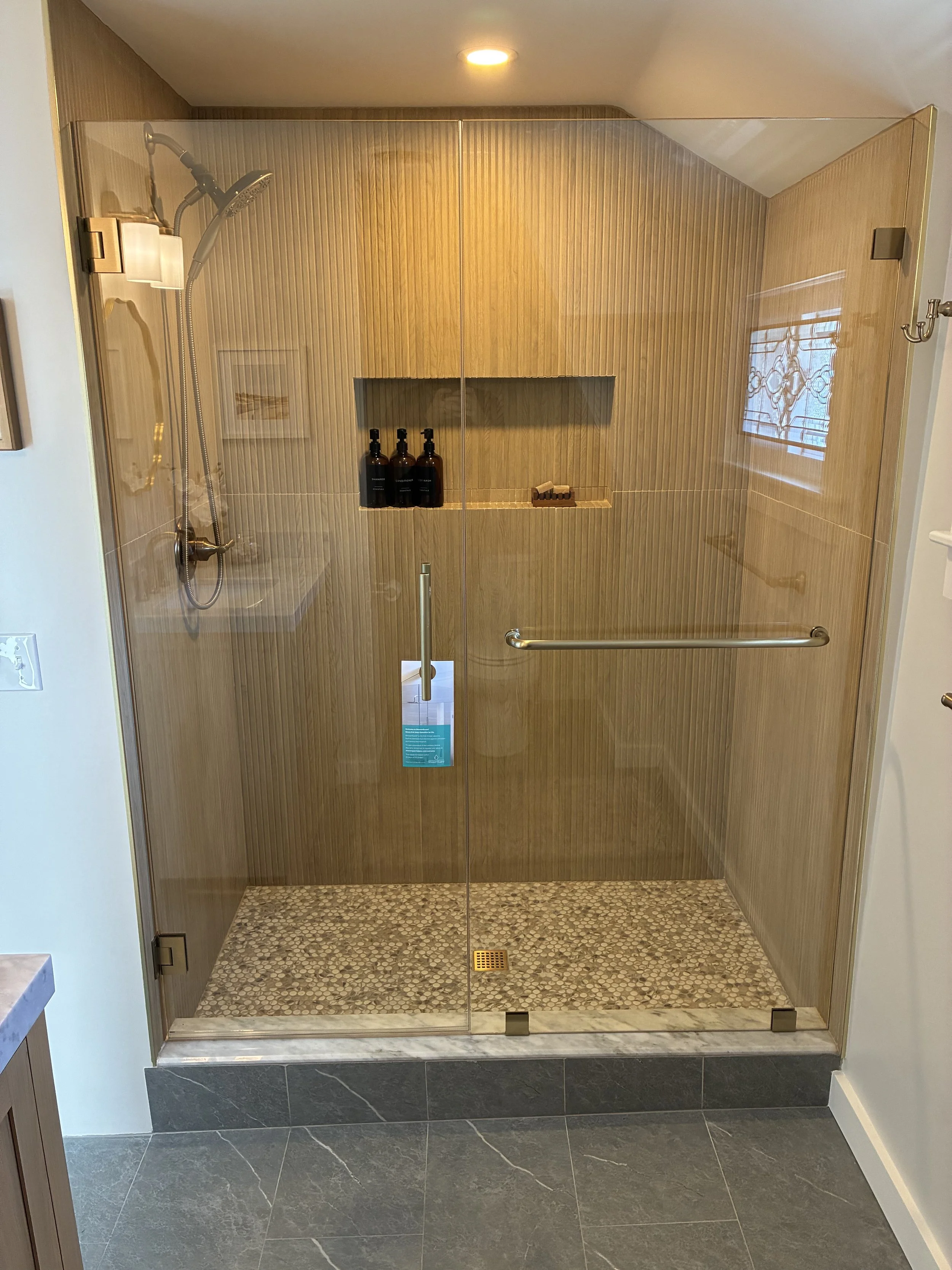 A modern glass-enclosed shower with wood-textured walls, a built-in shelf with three bottles and a bar of soap, and pebble-stone flooring, in a bathroom with gray tiled floor.