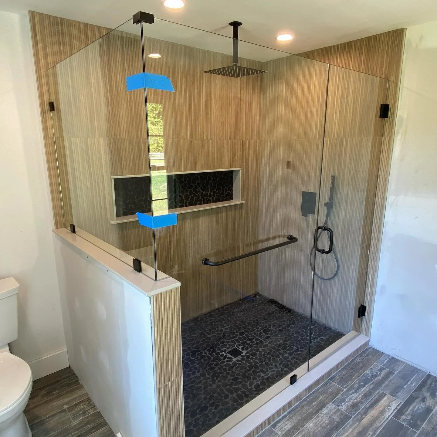 Frameless glass shower enclosure with a hinged door and a panel on a white knee wall, matte black hardware, wood-look tile walls, a long niche and a black pebble floor, in a Wells, Maine bathroom