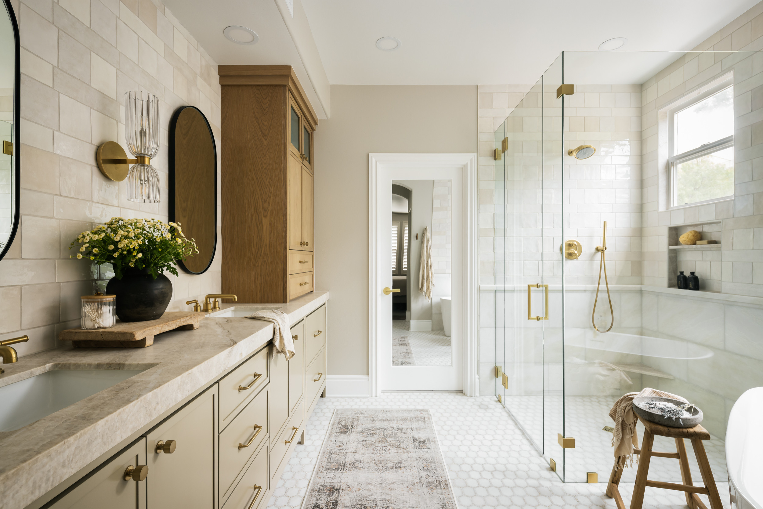 Modern bathroom with double vanity, white marble countertops, black oval mirrors, and brass fixtures. Tall wooden cabinet, patterned rug, and clear glass shower with brass accents. Small window, floral arrangement, and a stool with spa items.