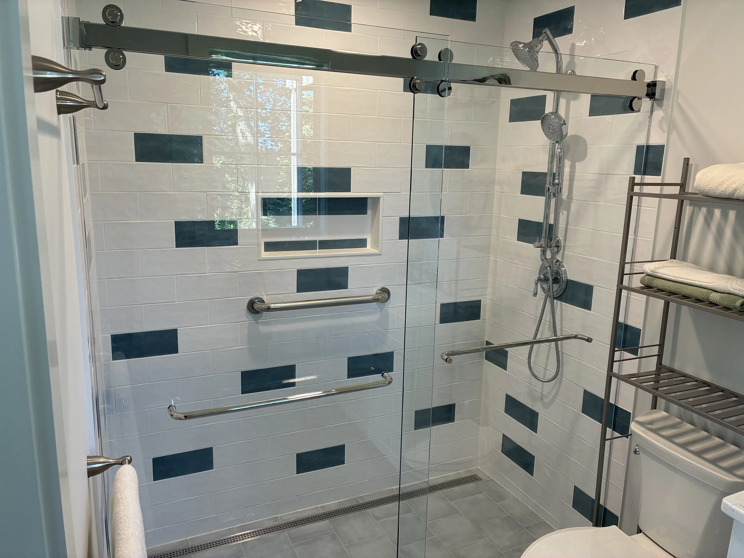Bathroom shower with glass door, white and dark tiles, and a built-in shelf. There are grab bars, a handheld showerhead, and a towel rack with towels. A shelving unit with towels and a toilet are visible.