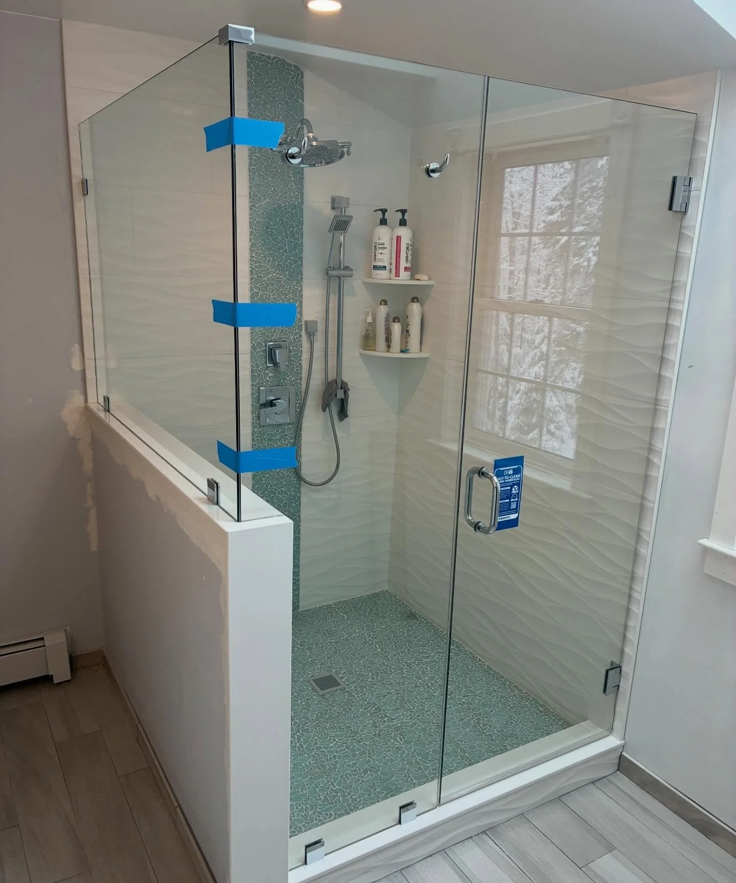 Frameless glass shower door and panel on a white knee wall, with chrome hardware, white wave tile, a glass mosaic stripe and a window, in a Scarborough, Maine bathroom