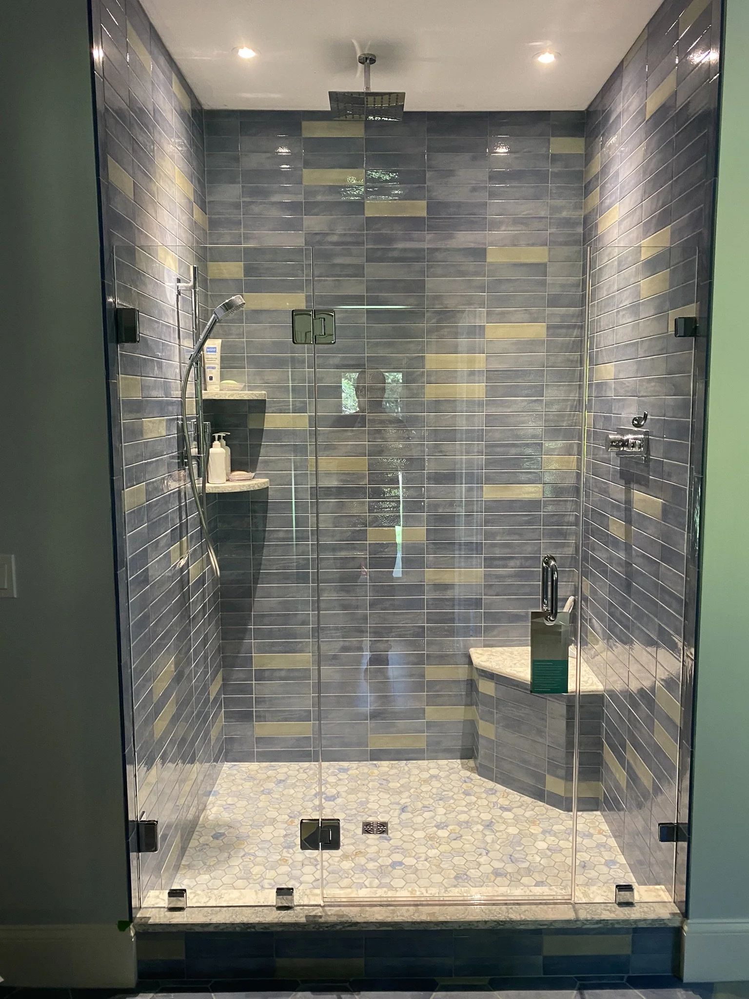 A glass-enclosed shower with blue and yellow tiles on the walls and small hexagonal tiles on the floor, featuring a rain showerhead on the ceiling, a handheld shower on the wall, a small seat, and shelves with toiletries.
