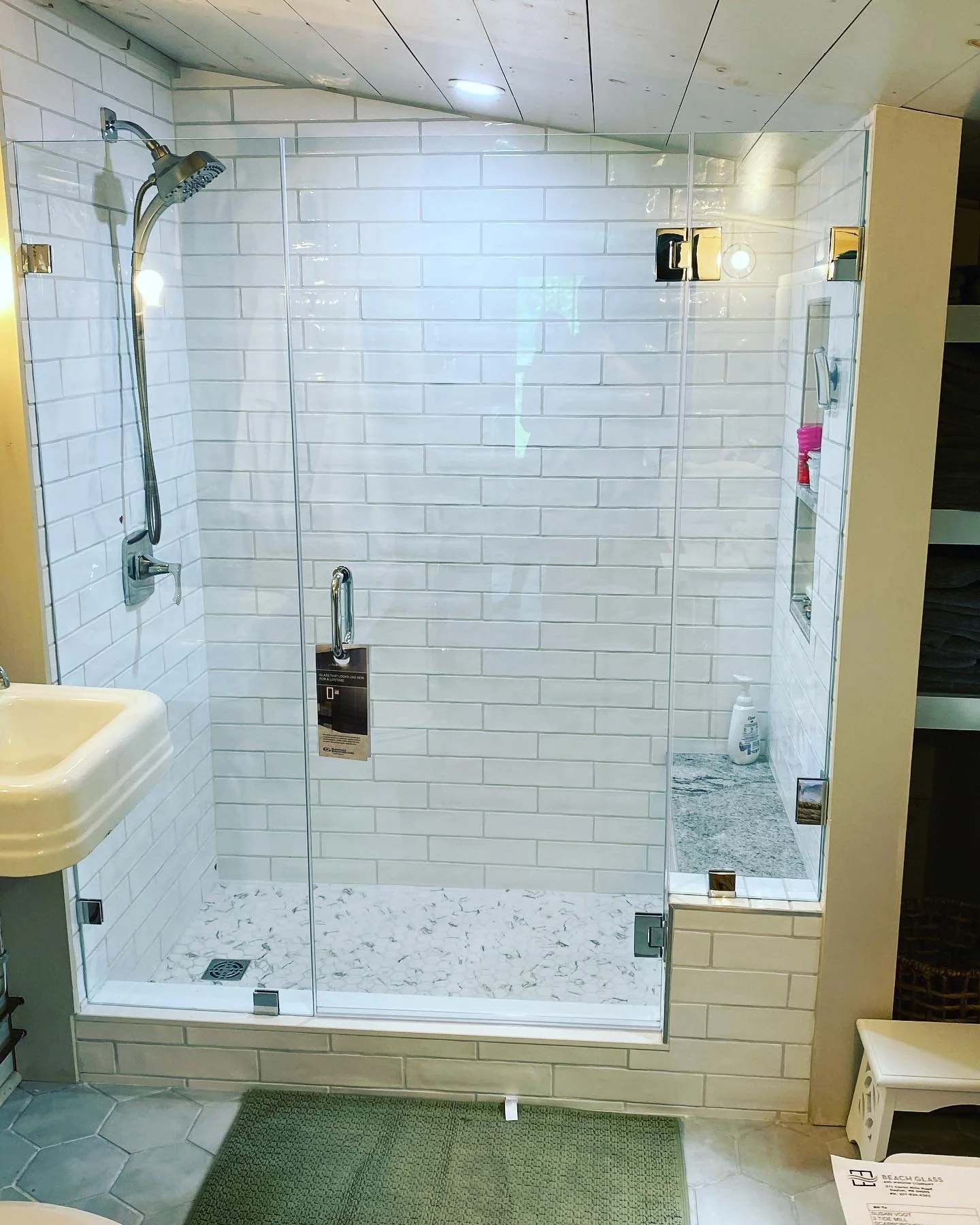 Frameless glass shower enclosure with a hinged door and panels on a tiled curb, white subway tile and a pebble floor, under a white plank ceiling in a Prouts Neck, Scarborough, Maine cottage bathroom