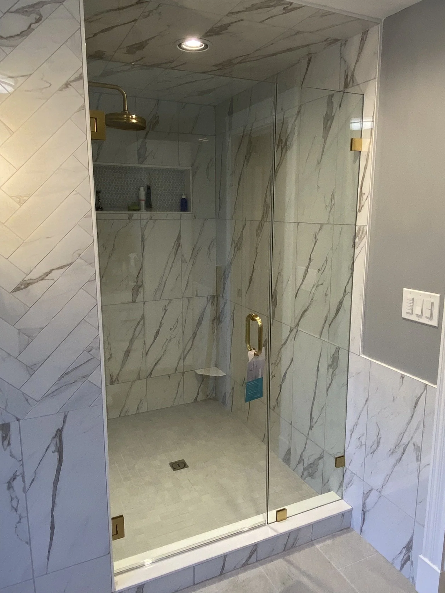 Bathroom shower with marble tiles, a brass shower head, glass doors, and built-in niche for toiletries.