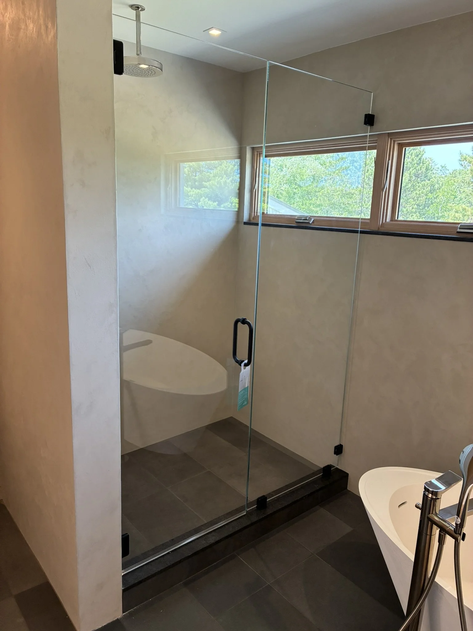 Modern bathroom with a glass walk-in shower, a window showing greenery outside, and a white bathtub with a chrome faucet.