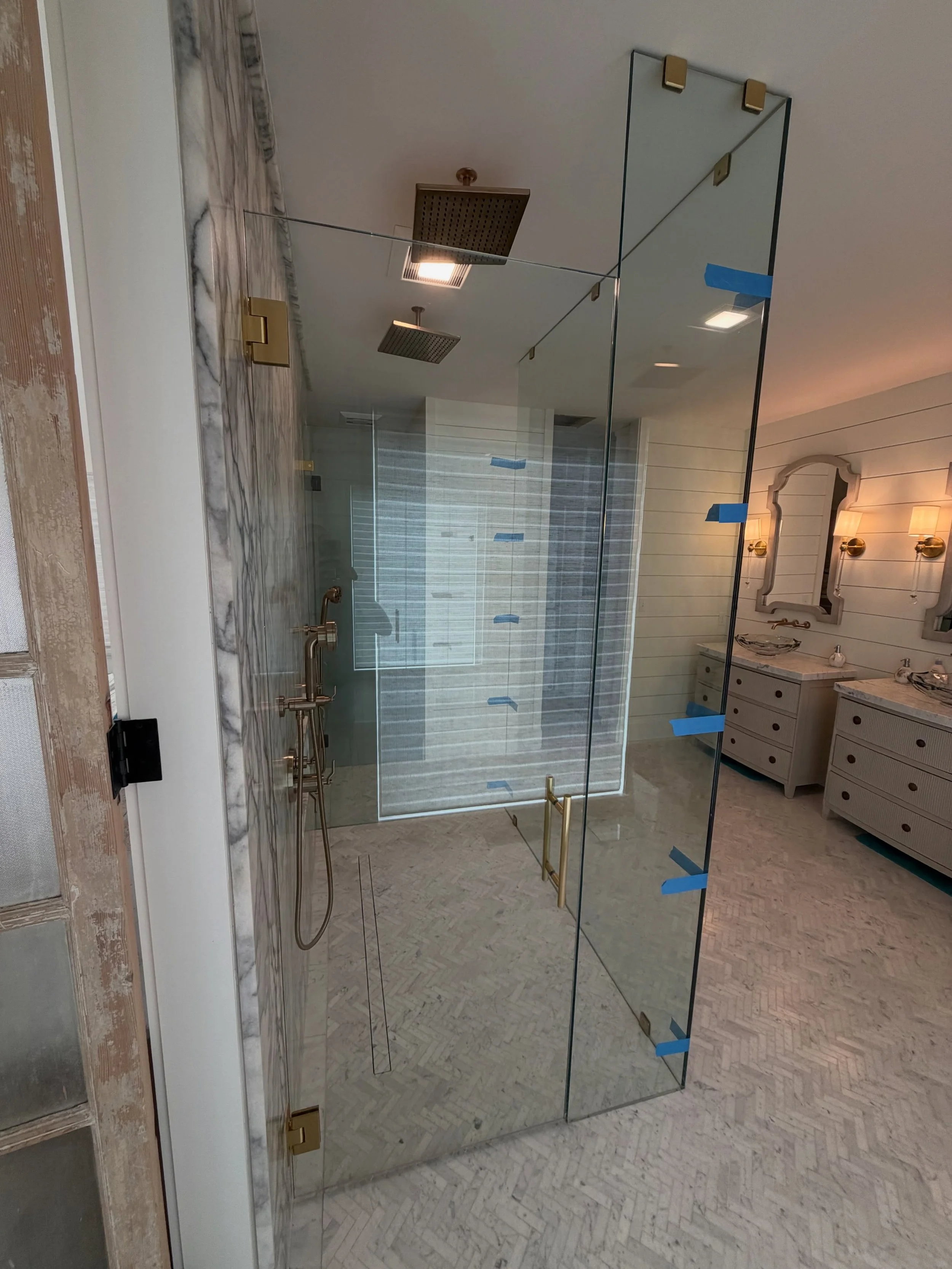 Tall frameless glass walk-in shower enclosure with brass hinges and clips, a ceiling rain head, blue and white tile and a marble herringbone floor, beside a double vanity in a Yarmouth, Maine bathroom