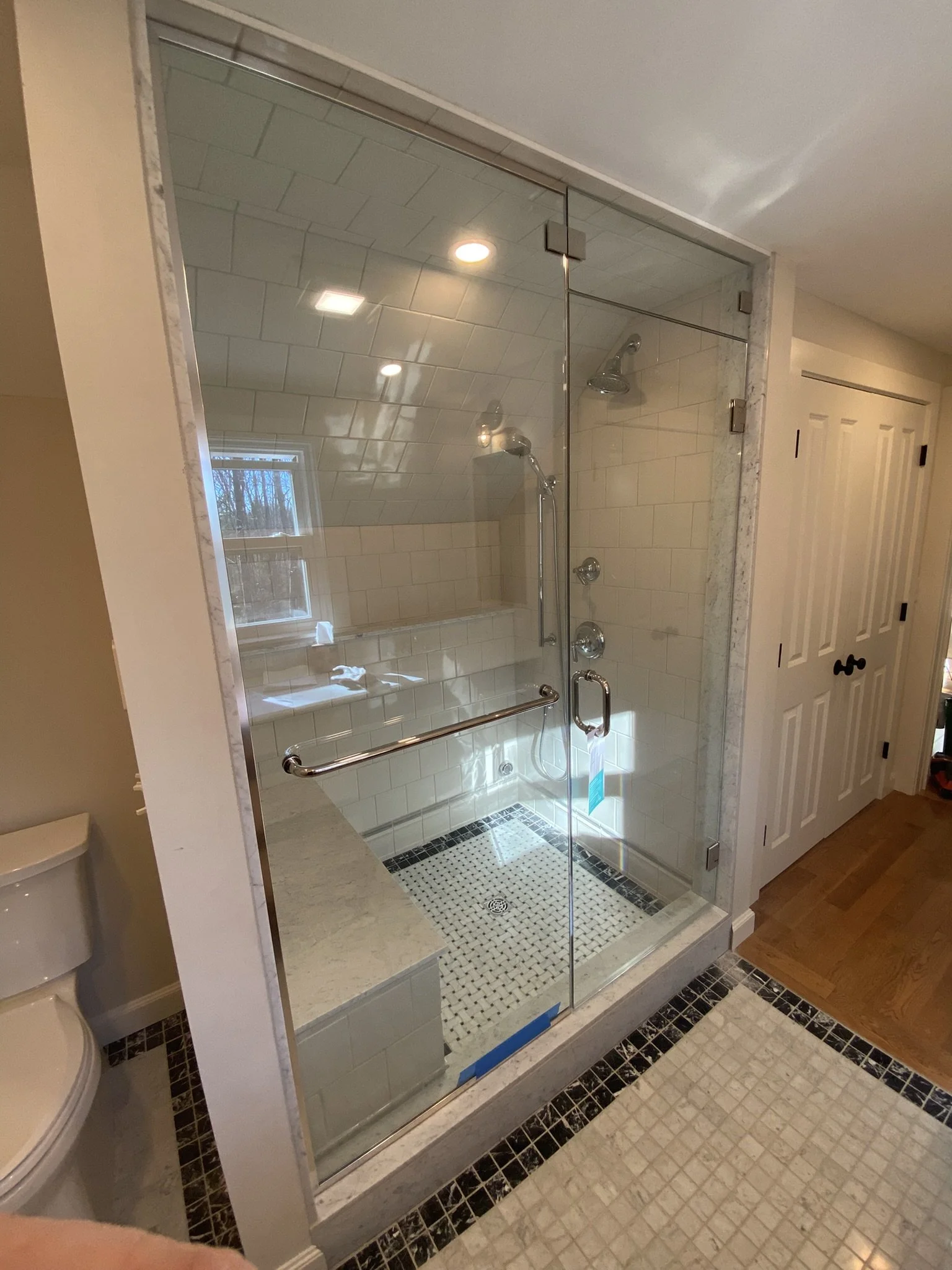 A glass-enclosed shower with white tiled walls and a built-in bench. The shower has two showerheads and a grab bar, and the floor has small black and white mosaic tiles. There is a small window in the shower area and a mirror reflecting the ceiling l