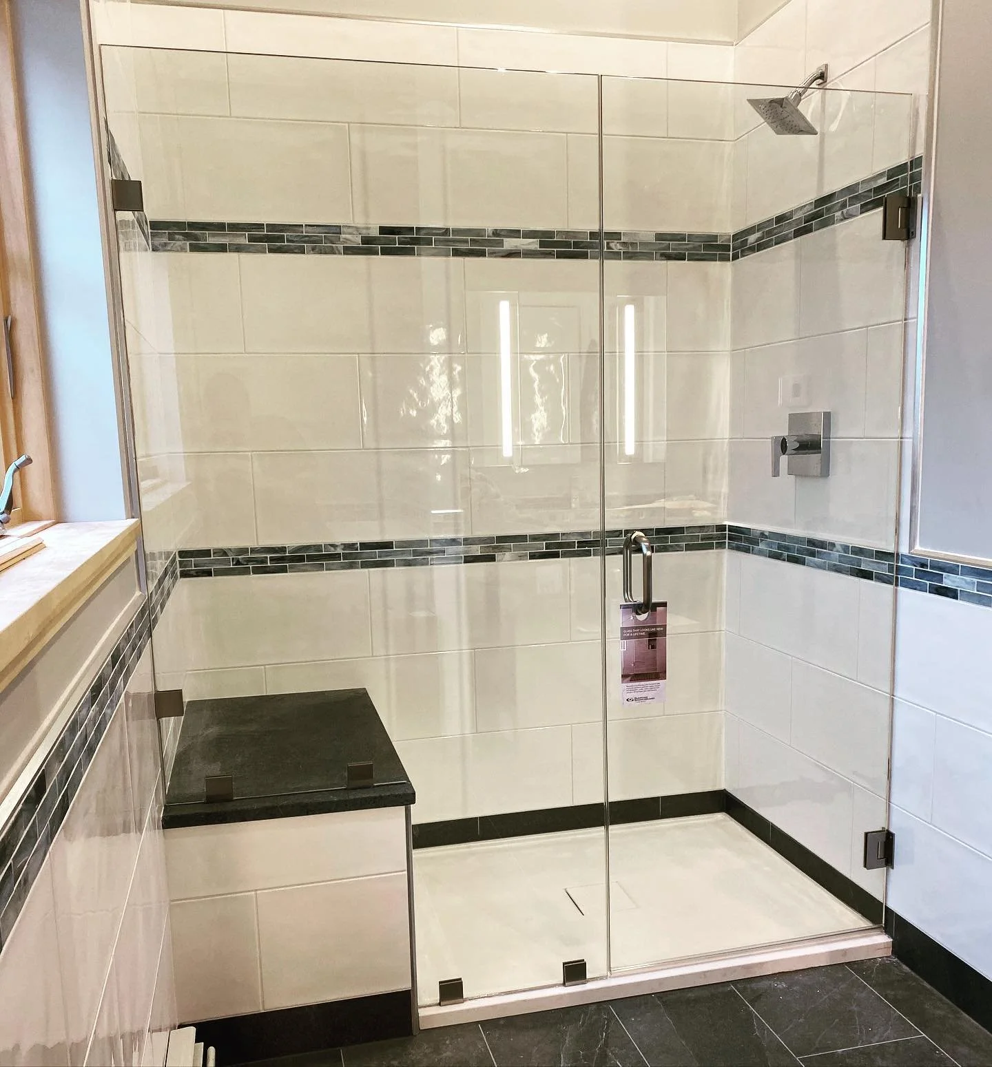 Frameless glass shower door and panel with chrome hinges, white tile with a dark glass mosaic band and a black stone bench, in a North Yarmouth, Maine bathroom