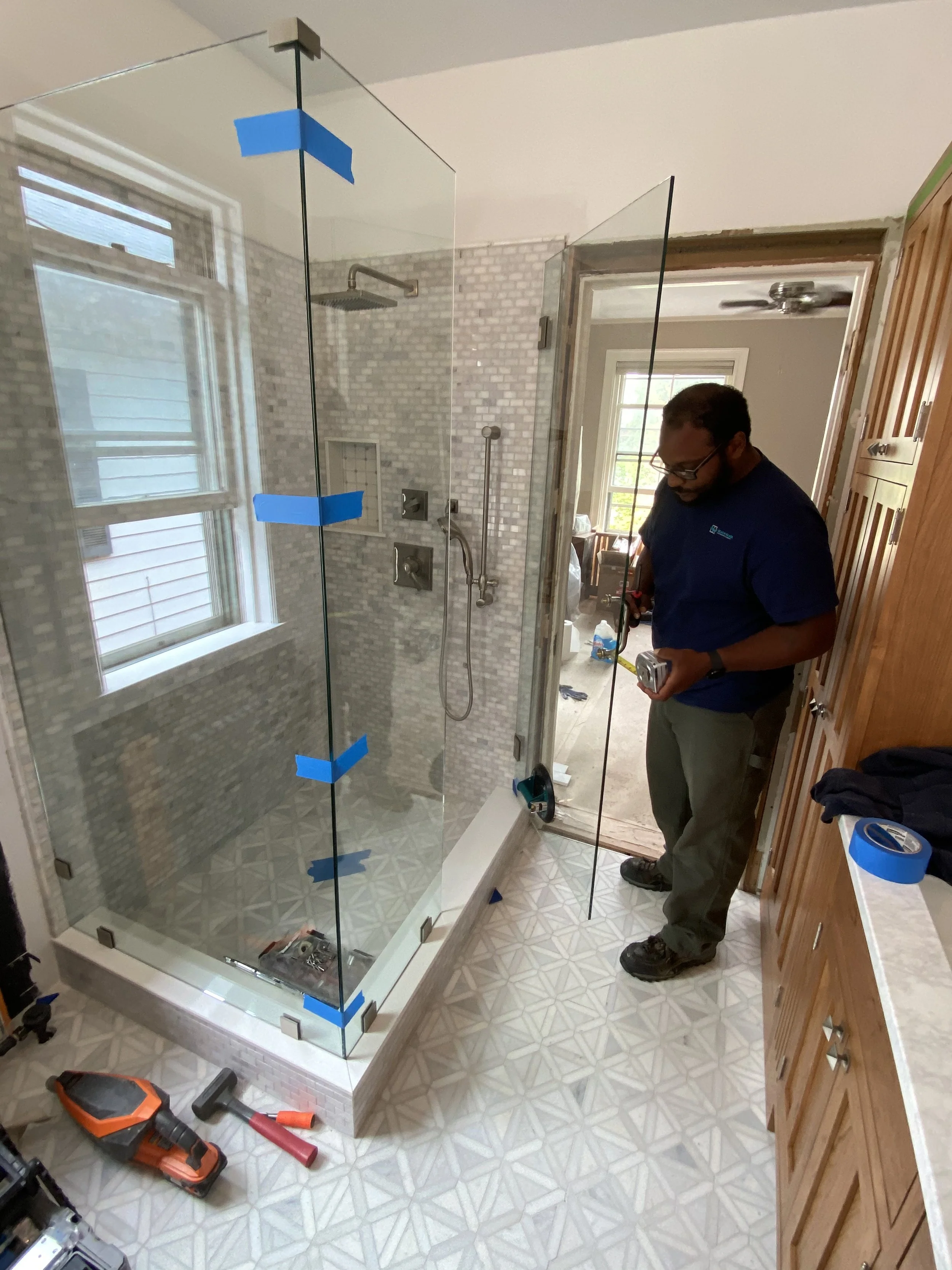 A man working on installing a glass shower enclosure in a bathroom. He is holding tools, and there are tools and blue painter's tape on the floor and counter. The bathroom has patterned tile flooring and a wooden cabinet.