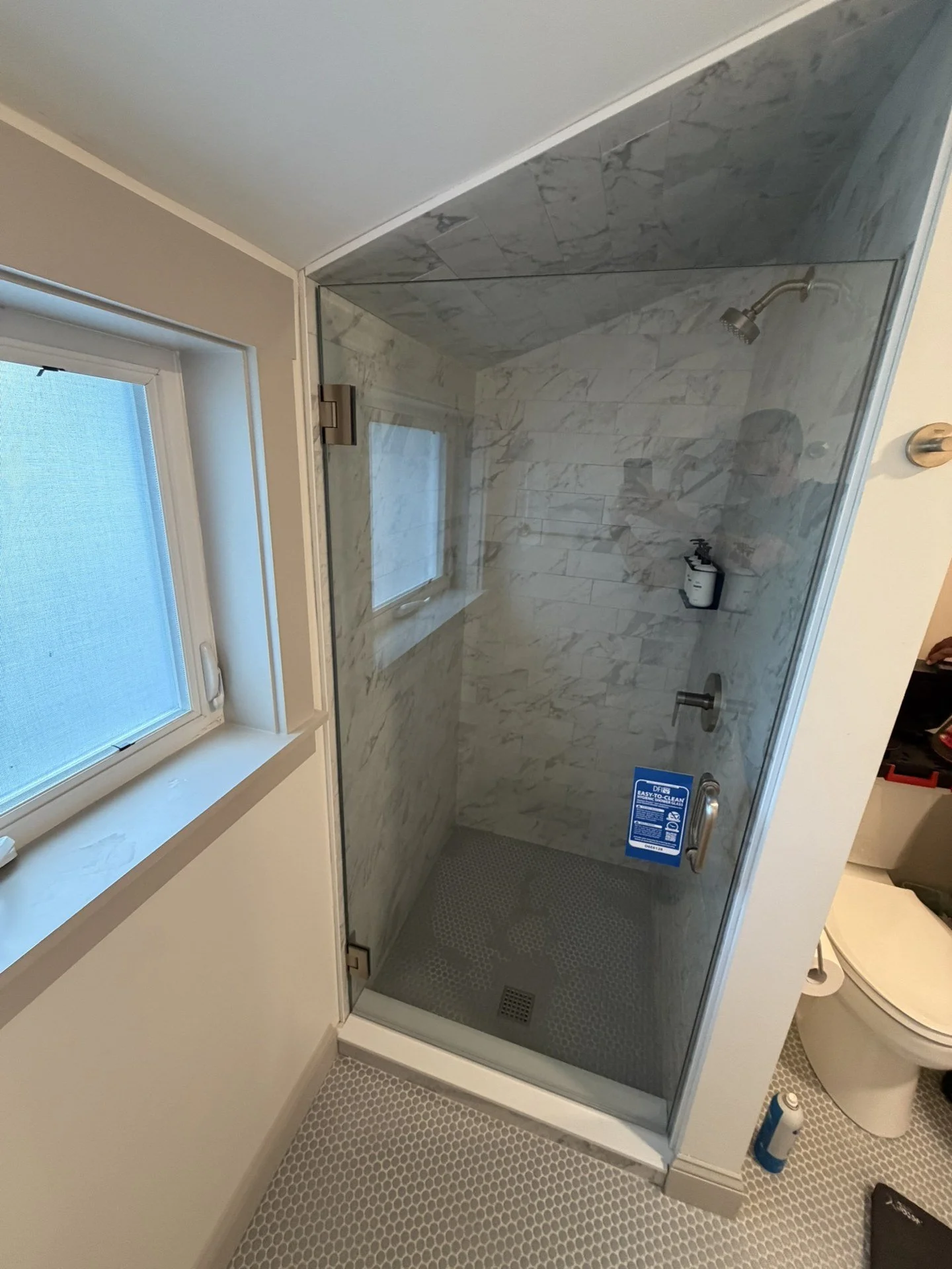 Frameless glass shower door with the glass cut to a sloped ceiling, marble-look tile walls and a white penny-tile floor, in a Yarmouth, Maine bathroom