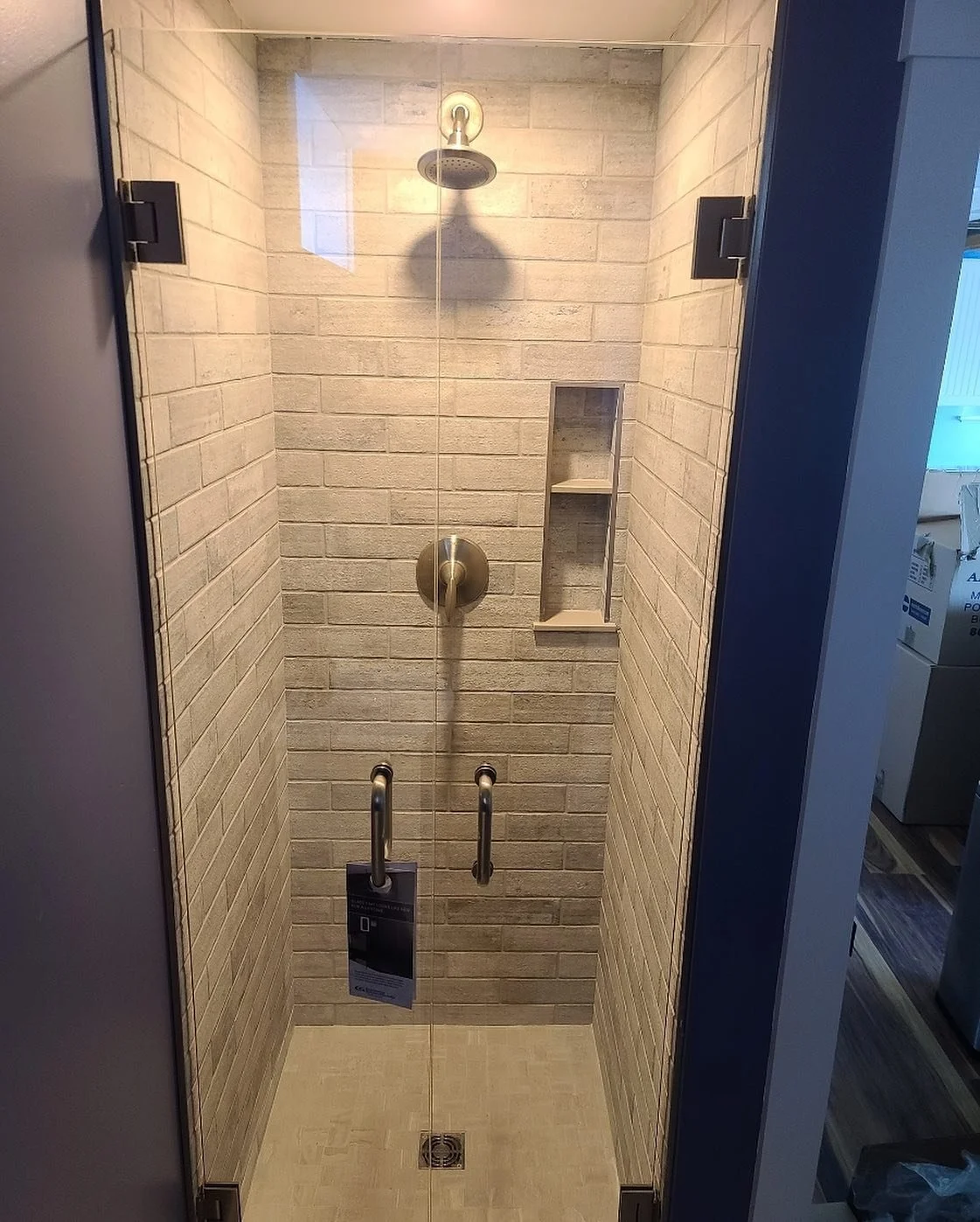 Pair of frameless glass shower doors opening from the middle, with matte black hinges, beige subway tile, a niche and brass fixtures, in a narrow Windham, Maine shower