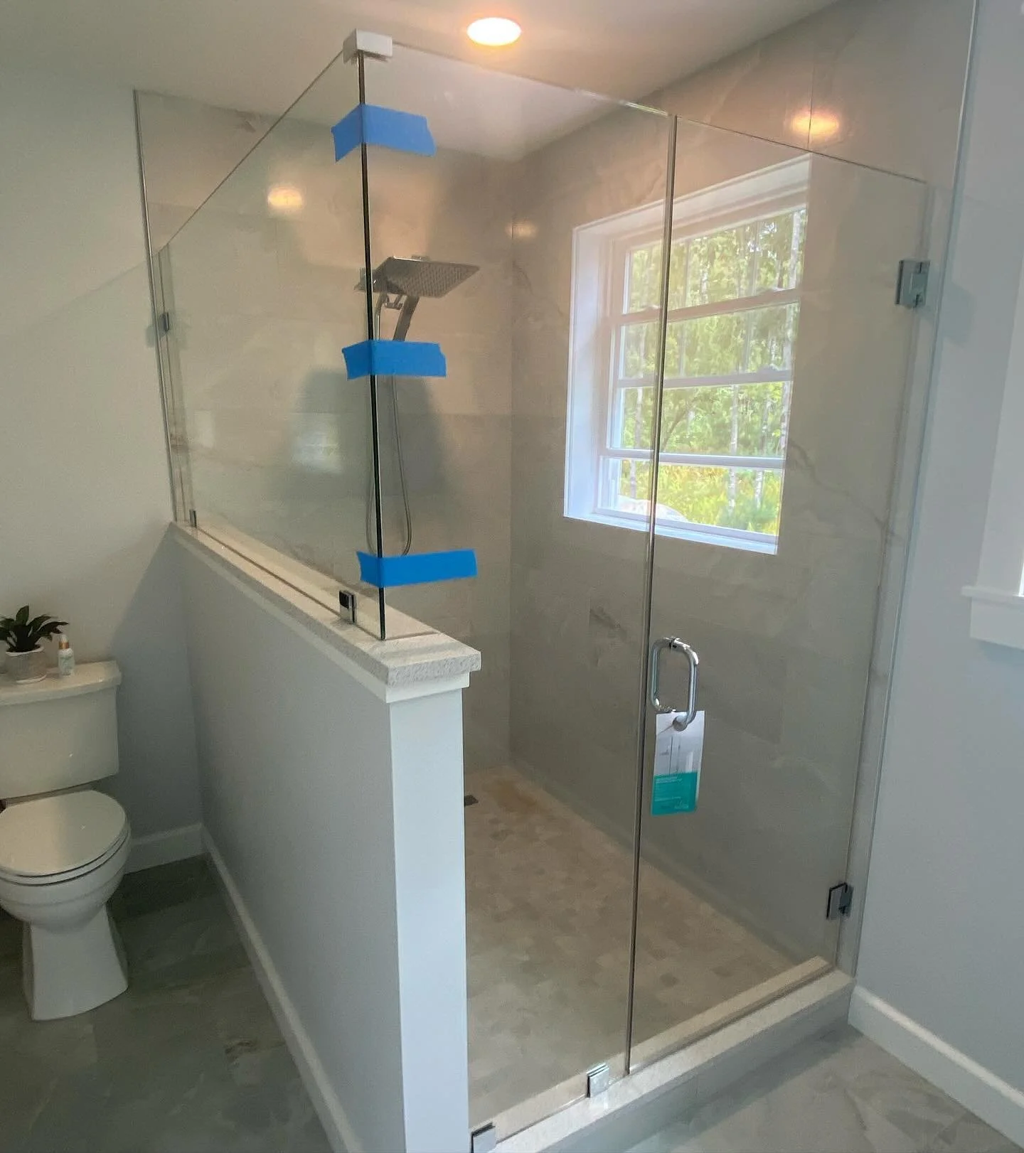 Large frameless glass shower enclosure with a hinged door and a panel on a white half wall, a window and gray tile, in a Hollis Center, Maine bathroom