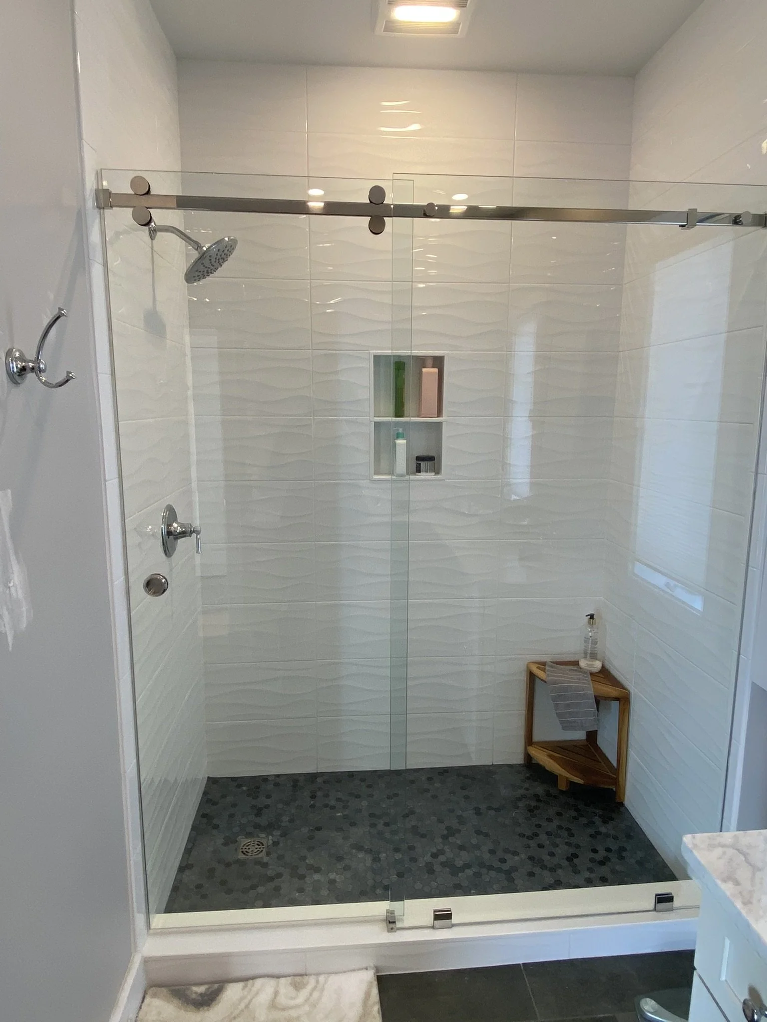 A modern walk-in shower with white textured wall tiles, a built-in shower niche with toiletries, a rainfall showerhead, a separate handheld shower, and a small wooden shelf with a water bottle and towel. The shower has a glass door and a square drain