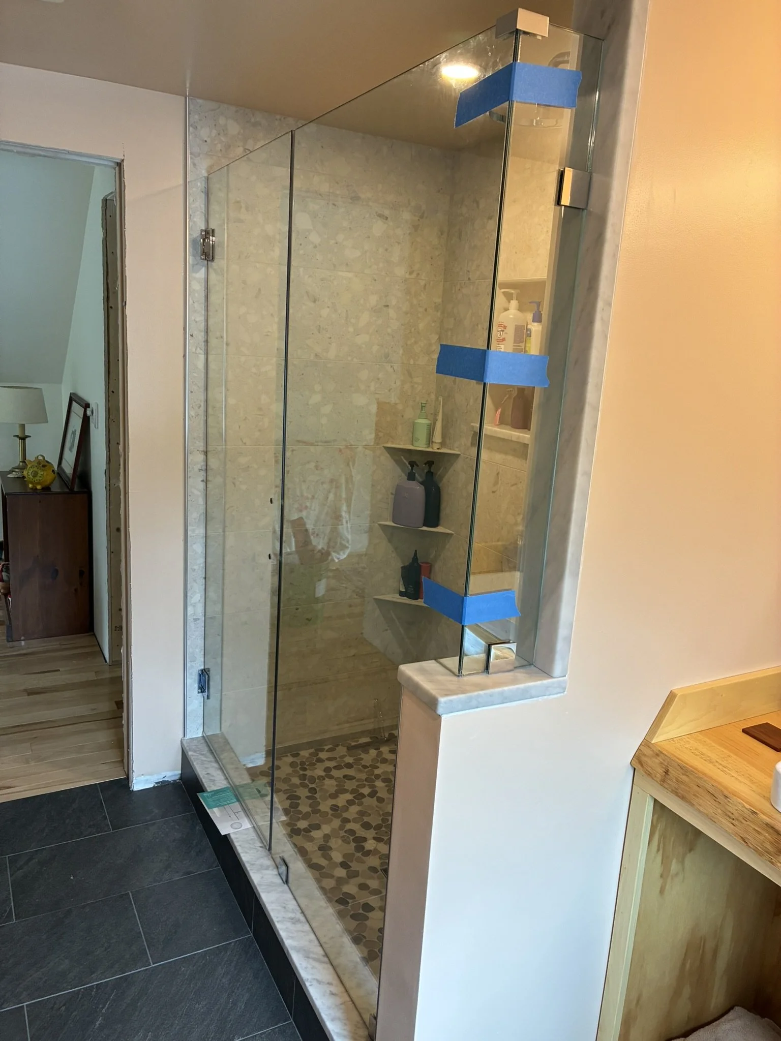 A glass shower enclosure with marble trim in a bathroom, containing three shelves with toiletries, and blue painter's tape on the edges of the glass.
