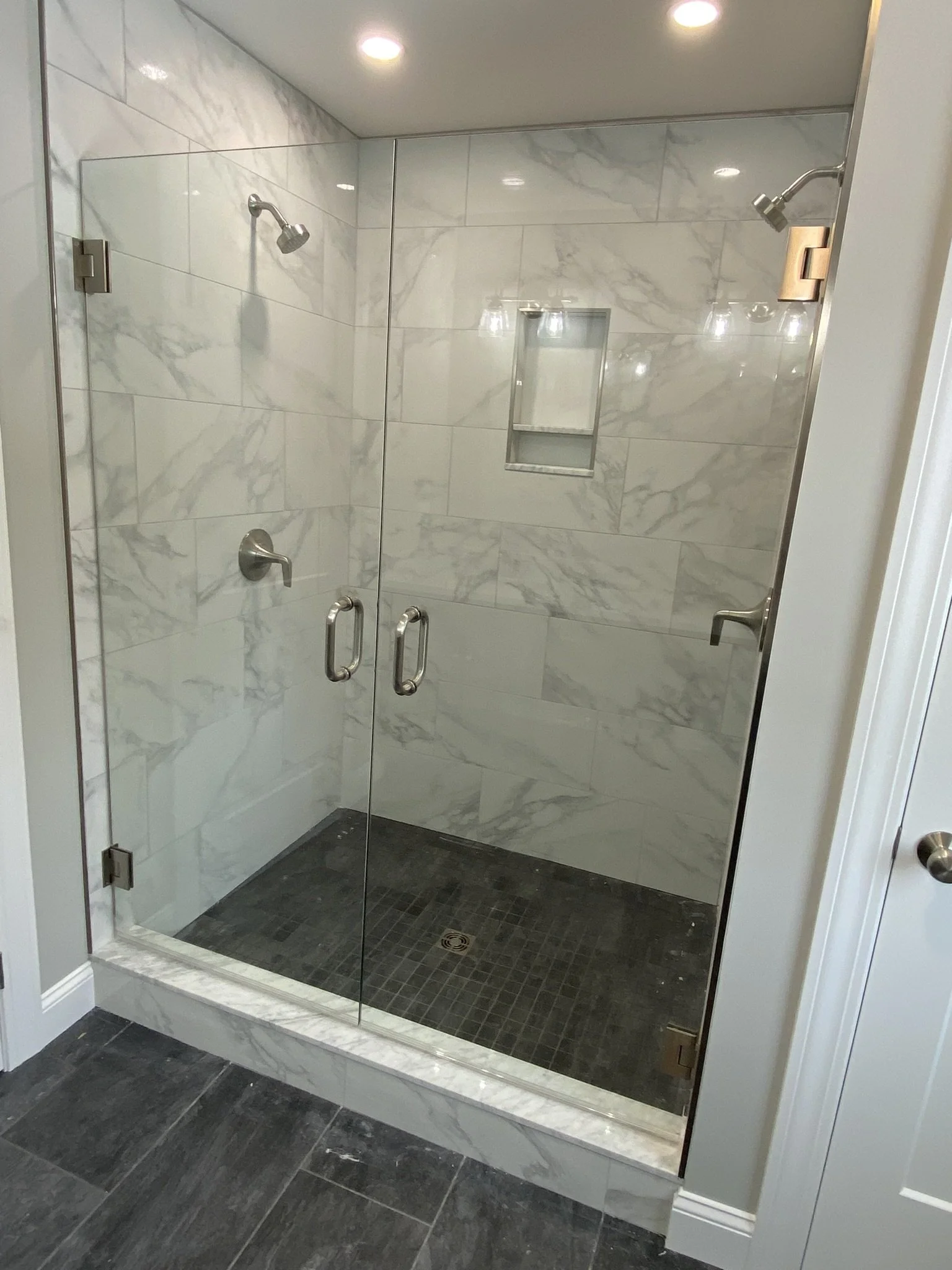 A glass-enclosed walk-in shower with marble tile walls, a built-in niche, a ceiling-mounted showerhead, and a dark tiled floor in a bathroom.