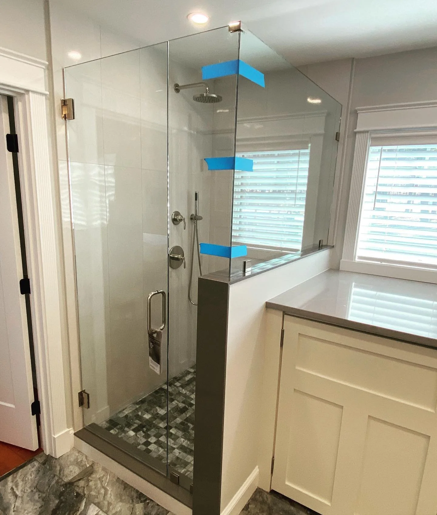 Frameless glass shower enclosure with a hinged door and a panel on a tiled knee wall, a checkered tile floor and a window, in a Sebago Lake, Maine bathroom