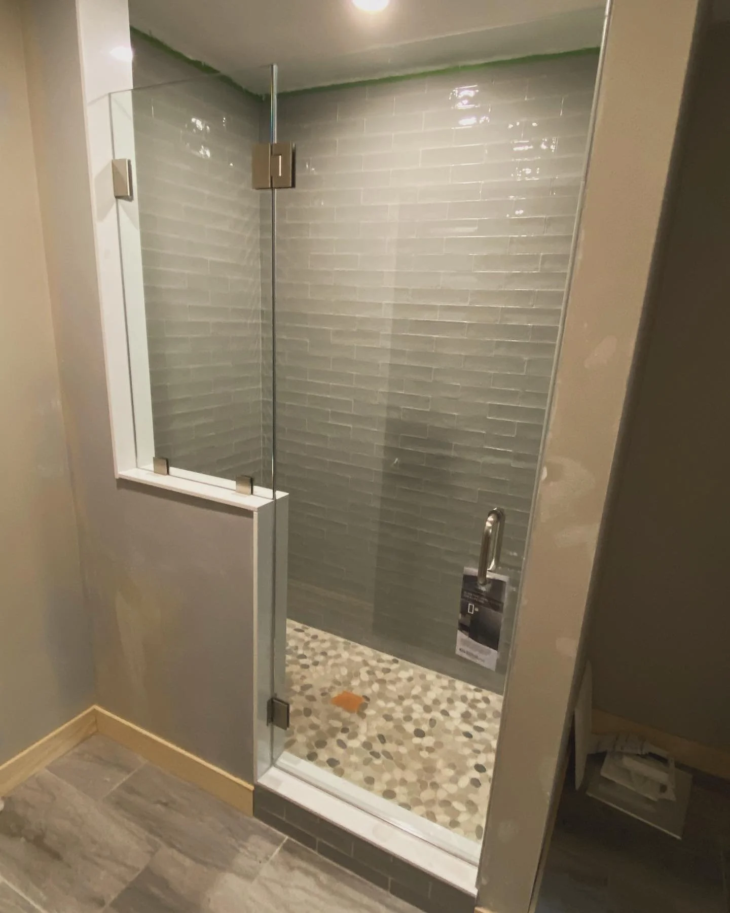 Frameless glass shower door and panel on a white knee wall, with chrome hinges, gray glass subway tile and a pebble floor, in a Kennebunk Pond, Lyman, Maine bathroom