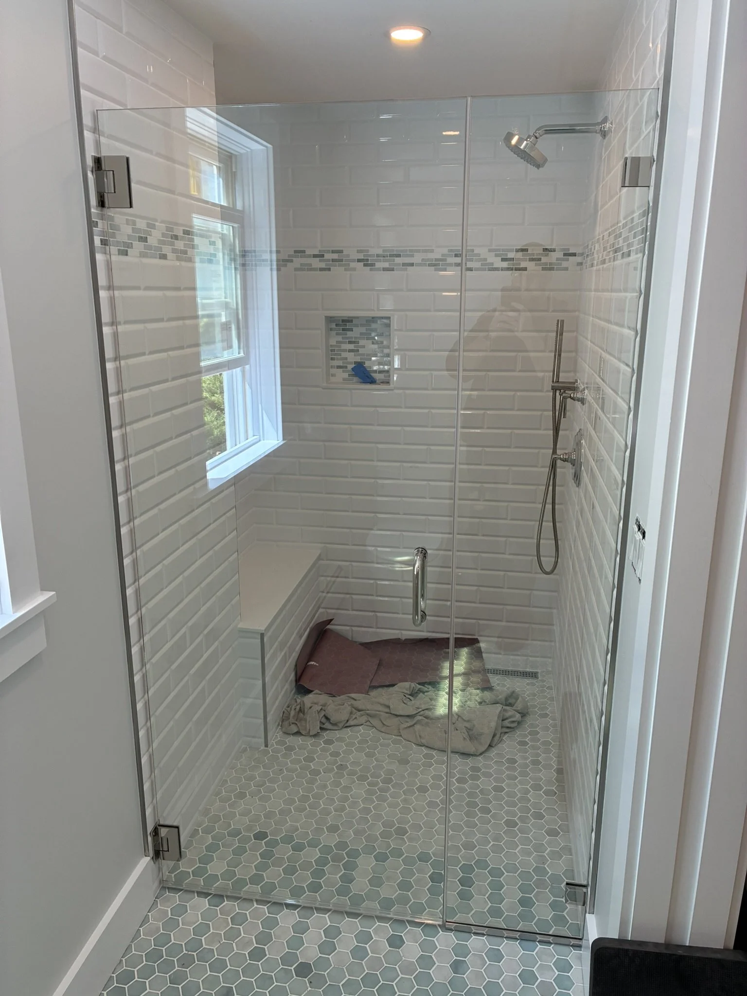 New shower with white subway tile on walls, a window, a built-in niche, hexagonal floor tiles, and a rain shower head with a handheld shower attachment. There are some towels or rags on the floor.