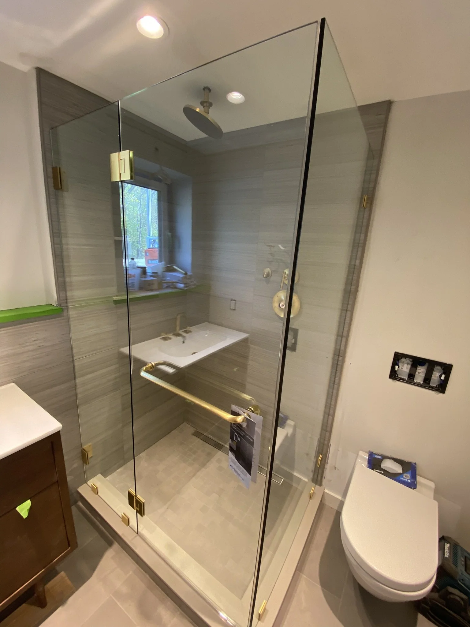 A bathroom with a glass shower enclosure, a window, and a toilet. The shower has a brass handle and fixtures, and the walls inside are tiled with grayish tiles. The toilet is white and modern, and there is a small cabinet visible to the left and a wa