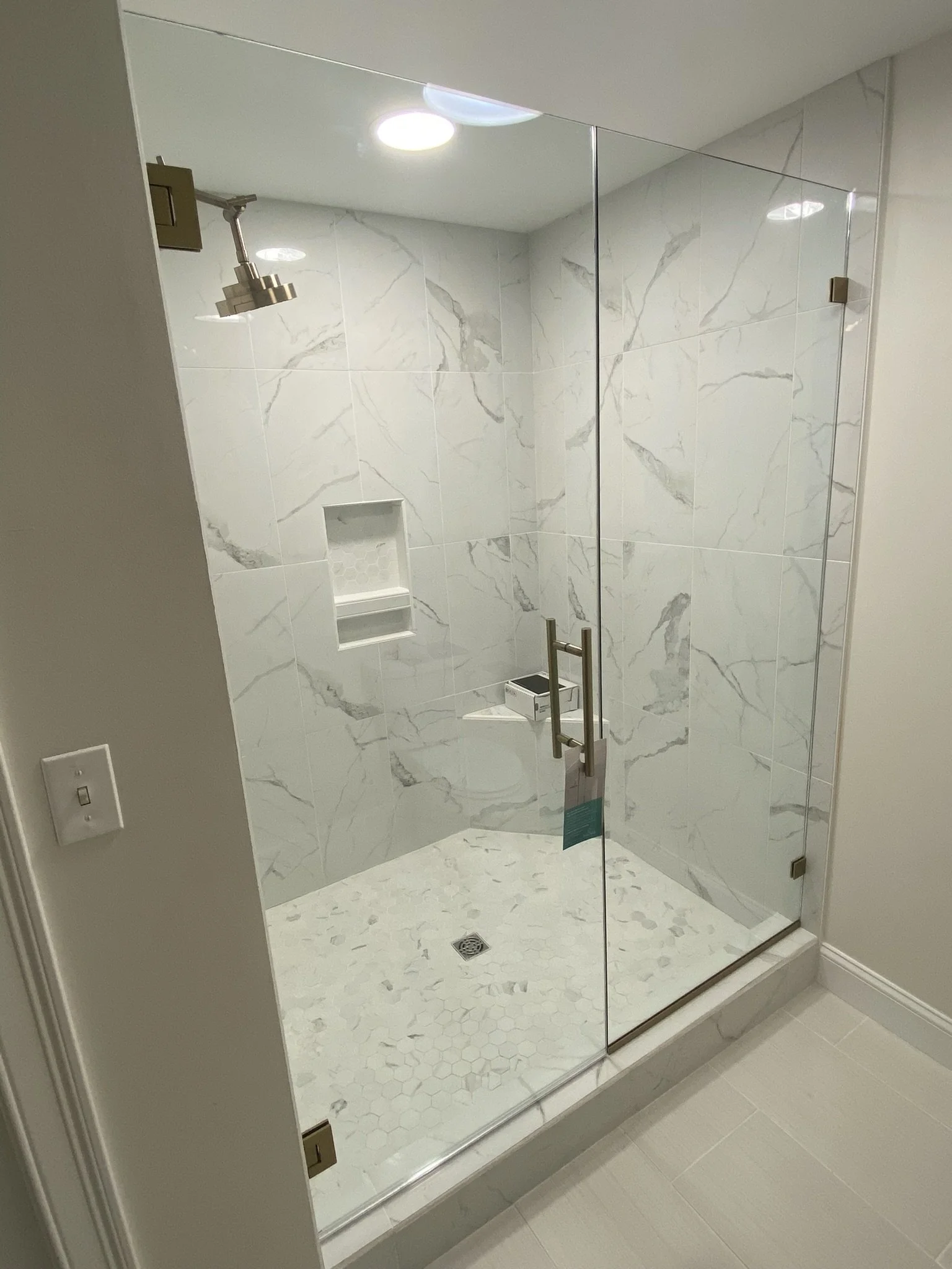 A modern glass-enclosed shower with white marble tiles on the walls and a white hexagonal tile floor. There is a built-in niche for toiletries, a showerhead on the left wall, and a door handle on the glass door.