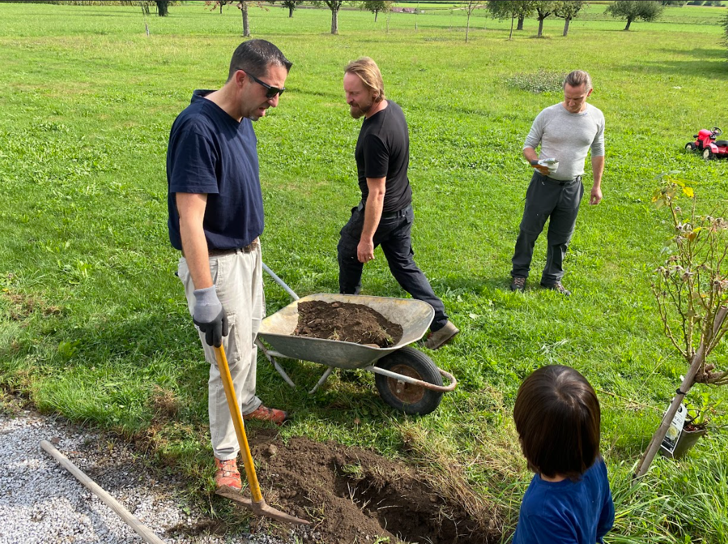 Menschen arbeiten im Garten, umgeben von grüner Wiese und Bäumen, mit Schubkarre und Gartenwerkzeugen.