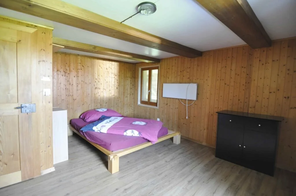 Schlafzimmer mit Holzwänden, einem Bett mit pinker Bettwäsche, einem kleinen Fenster, einem schwarzen Schrank und einer weißen Wandheizung.