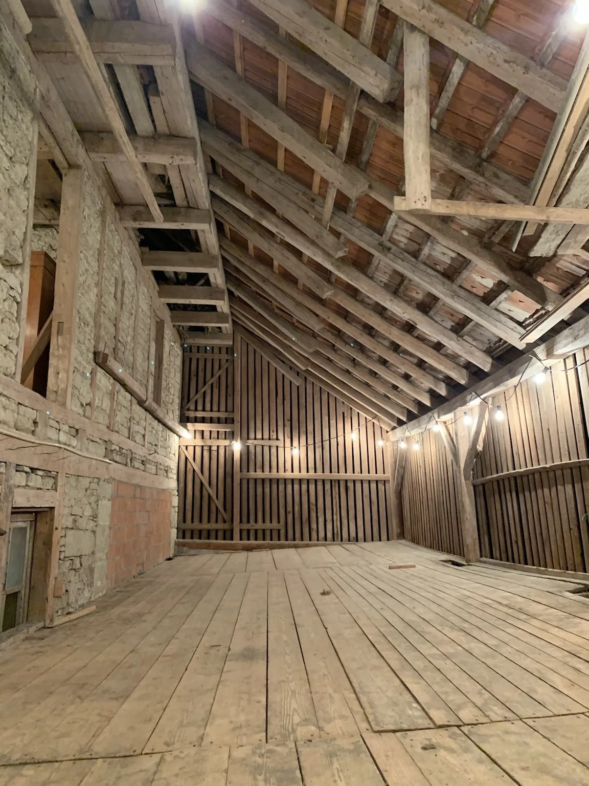 Innenraum einer unbeheizten, halbrenovierten Scheune oder Abstellraum mit Holzboden, Holzwänden und einem Holzschindeldach, beleuchtet mit einer Lichterkette.
