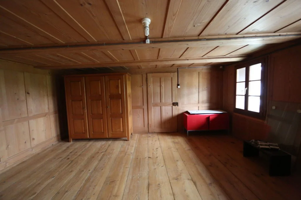 Ein leeres, rustikales Zimmer mit Holzvertäfelung an Wänden, Decke und Boden, ein brauner Kleiderschrank, eine rote Kommode, ein Fenster, einige Gegenstände auf einem schwarzen Kasten.