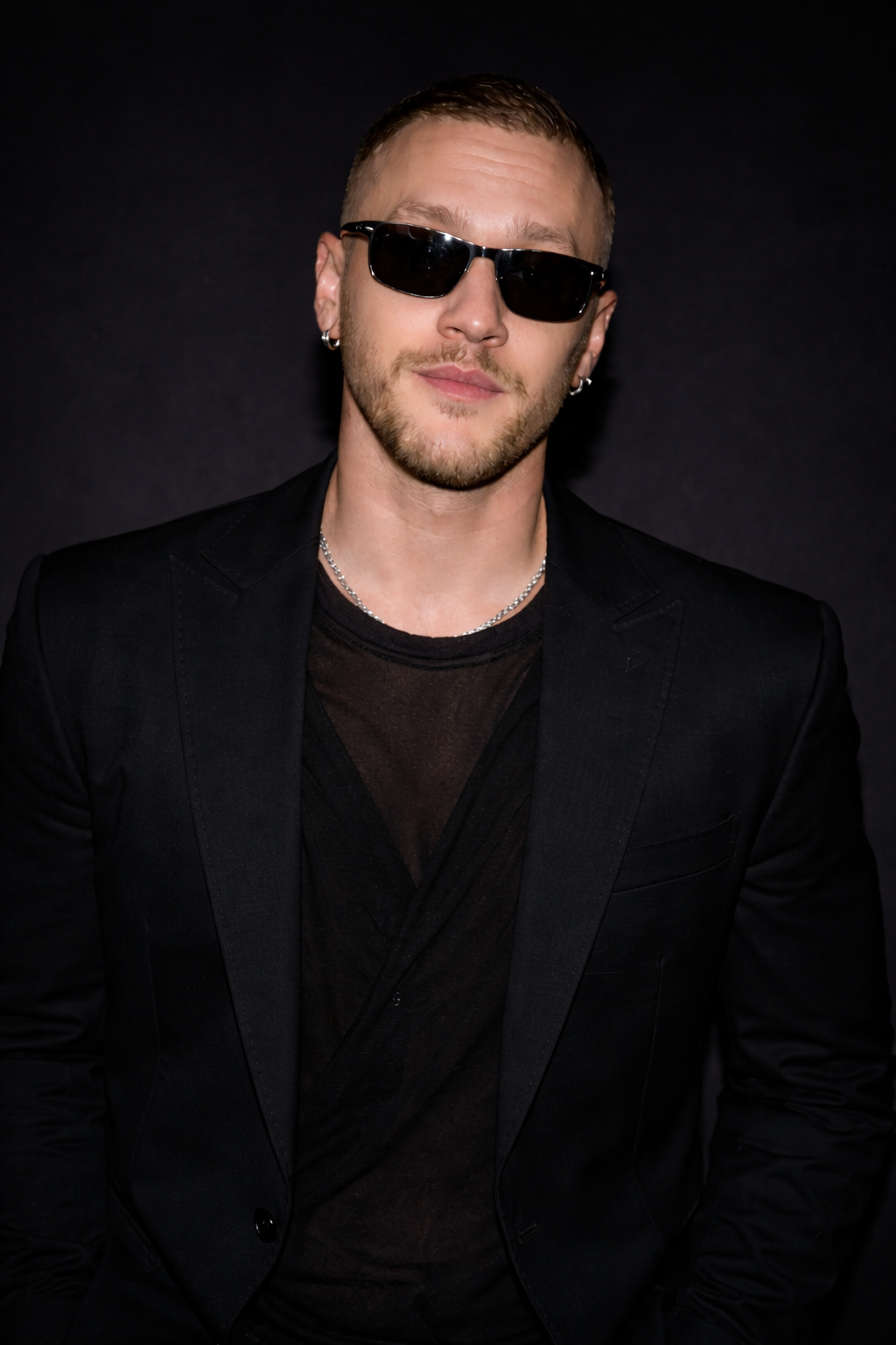 A self portrait of Govner.  A man with short hair, beard, and sunglasses wearing a black blazer, black shirt, and silver jewelry against a dark background.
