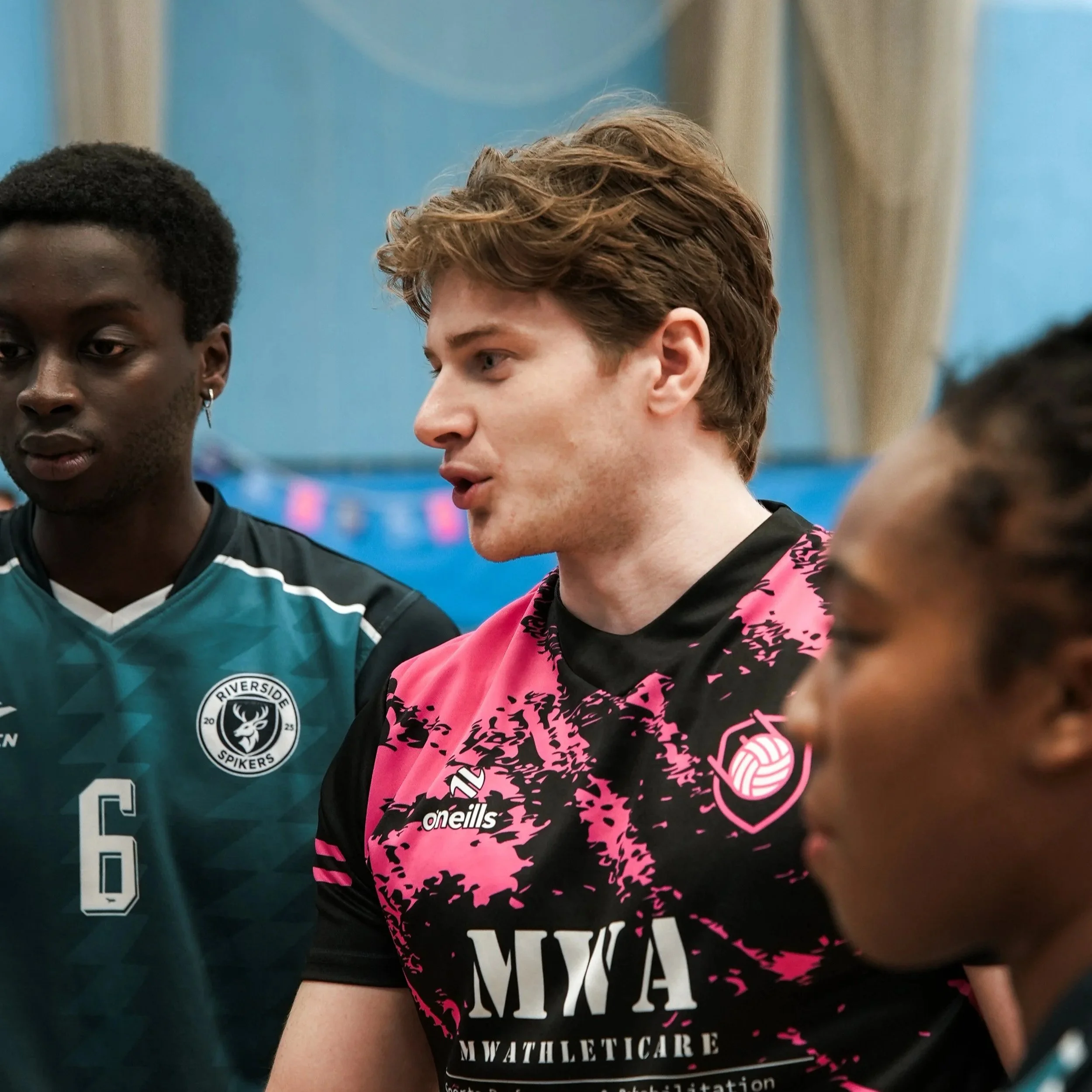 Close-up of a group of young athletes in sports jerseys, engaged in a conversation during practice or a game, with a focus on a young man with light brown hair wearing a black and pink jersey.