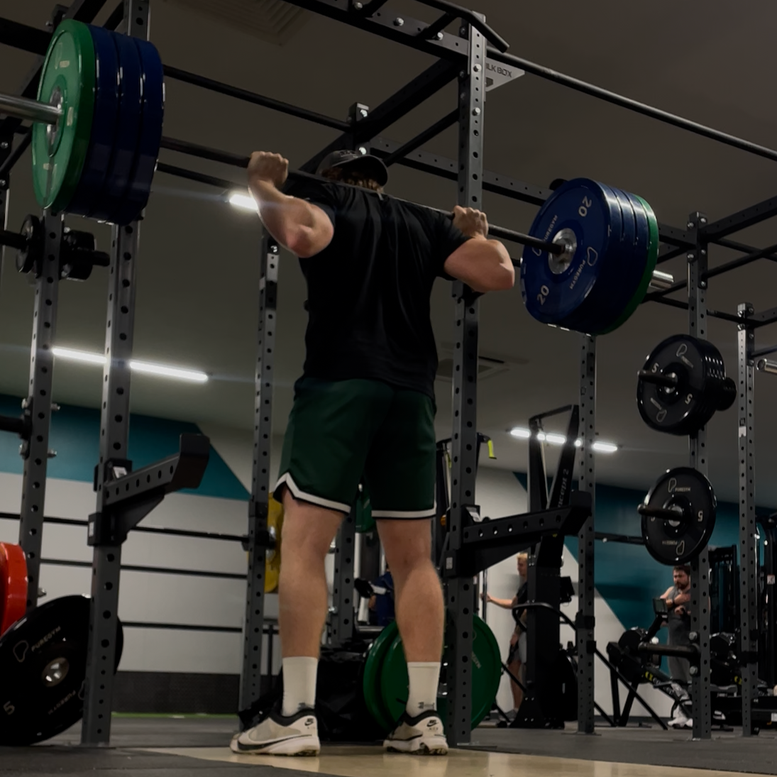 A man performing a back squat exercise with a loaded barbell in a gym, surrounded by weight plates and gym equipment.