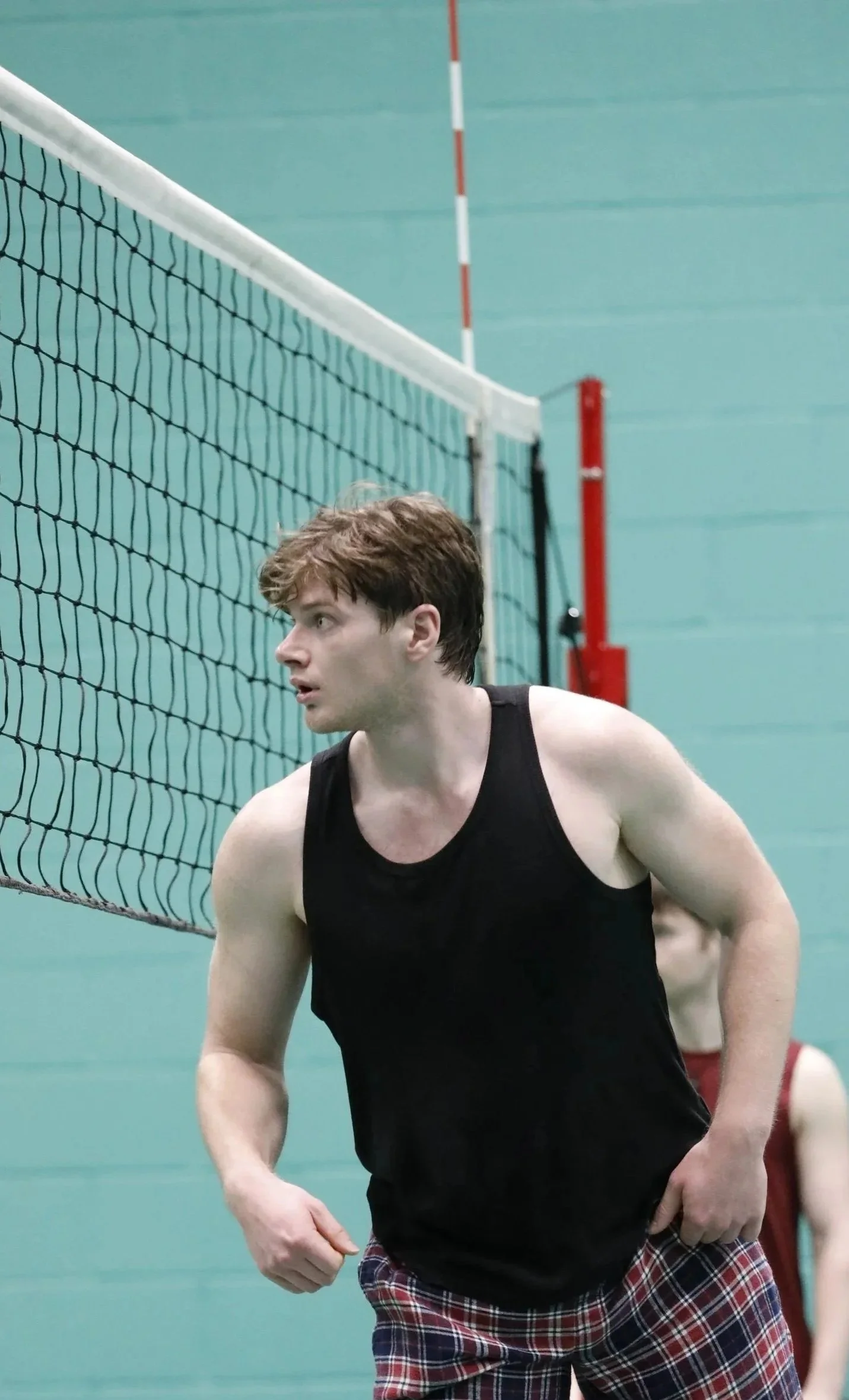 A young man with short brown hair, wearing a black sleeveless shirt and red plaid shorts, playing volleyball indoors.