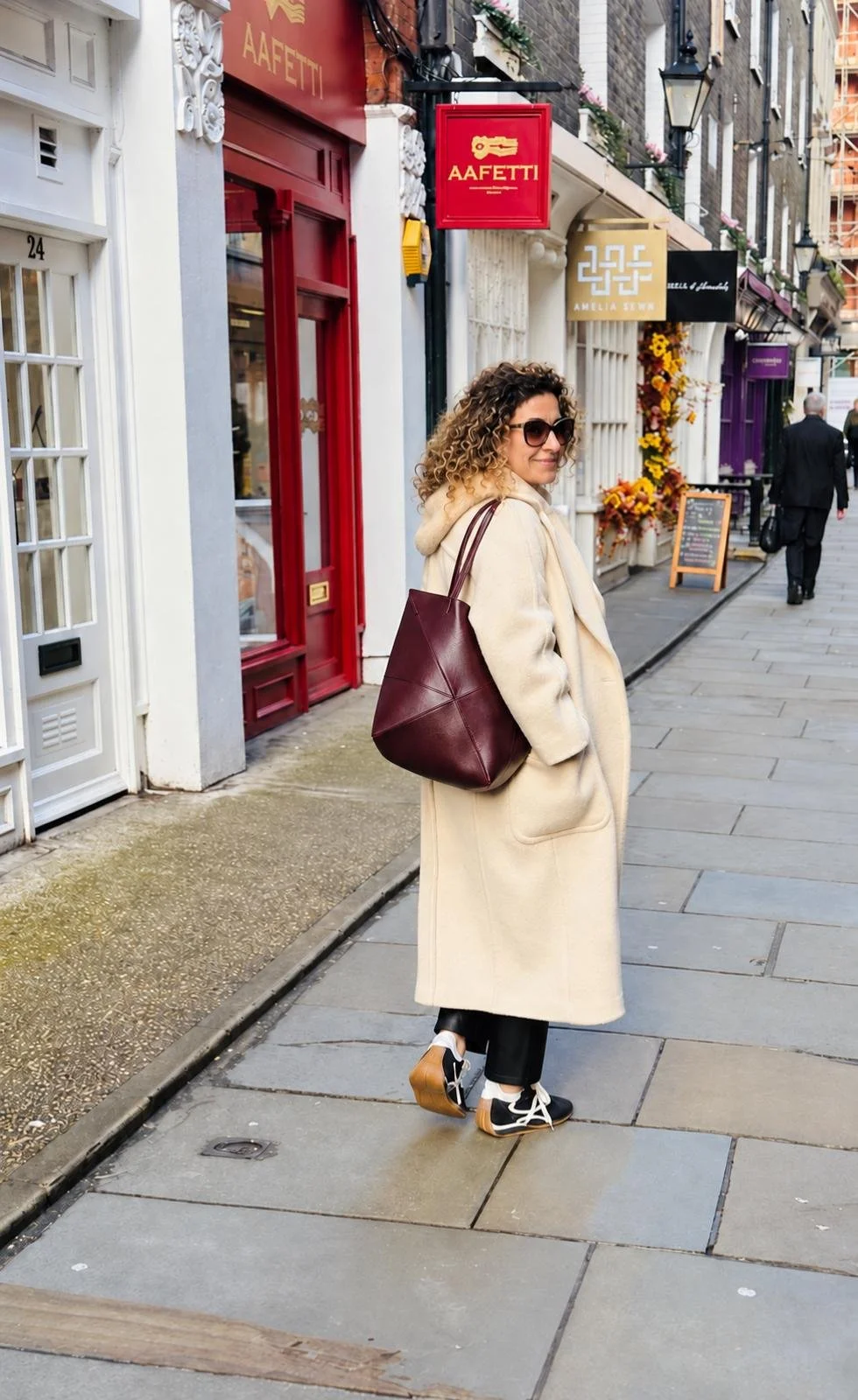 Alice , a personal stylist, shopping in central London with curly hair wearing sunglasses, a beige coat, black pants, and sneakers, standing on a city sidewalk with shops and signs behind her, carrying a large maroon handbag.