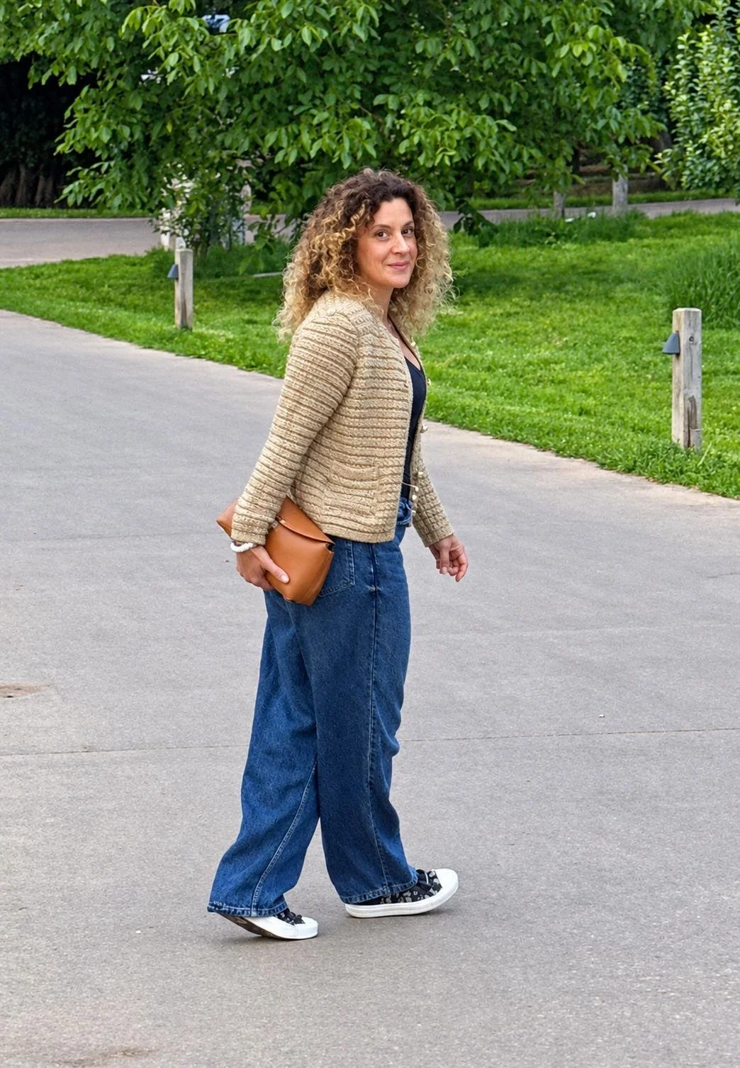 Alice, a personal stylist, dressed casually. A woman with curly hair dressed in a beige textured jacket, black top, baggy blue jeans, and black sneakers with white soles walking on a paved path in a park.