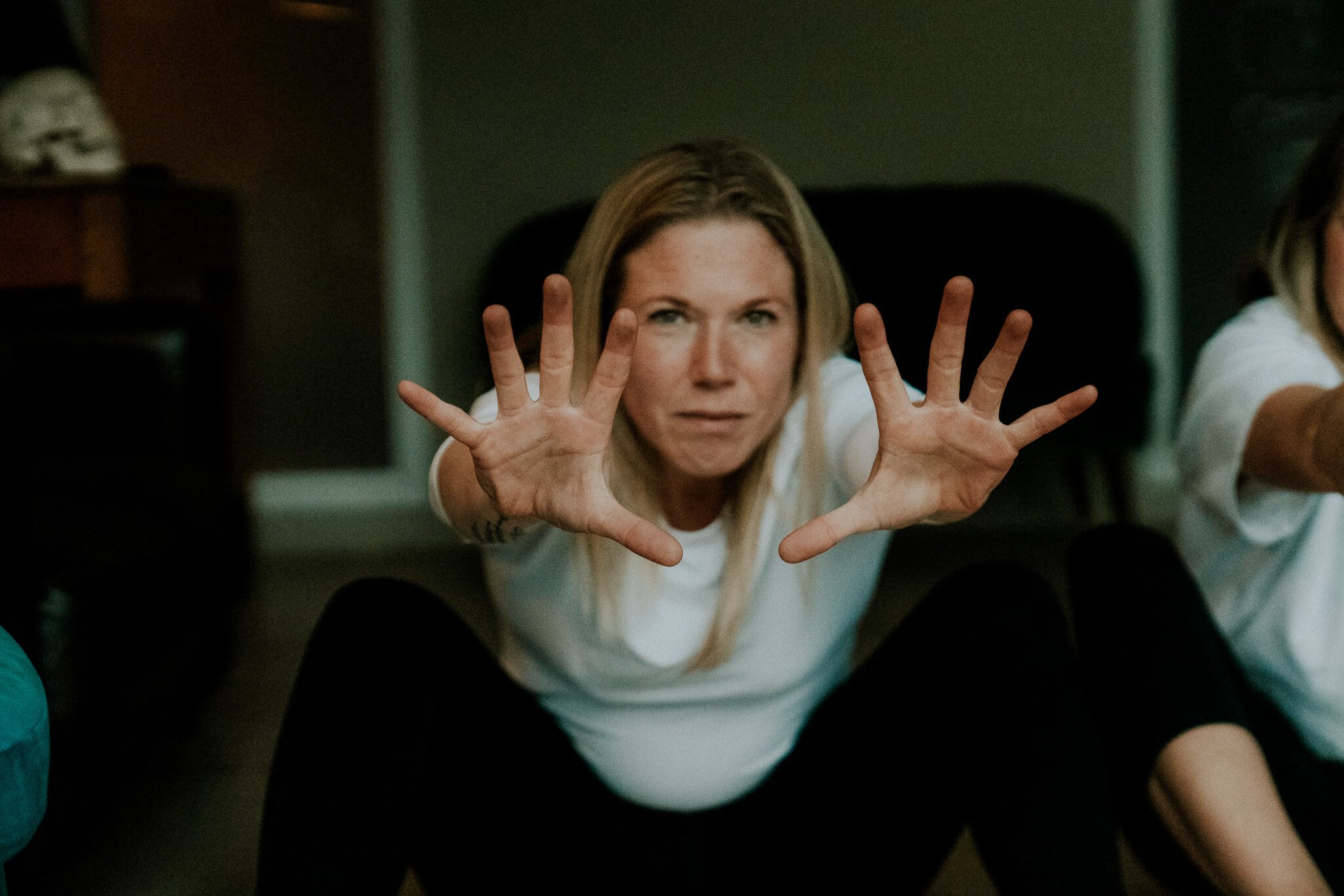 Abi Adams is sitting on the floor, bent forward with her arms stretched forward and towards the camera.
