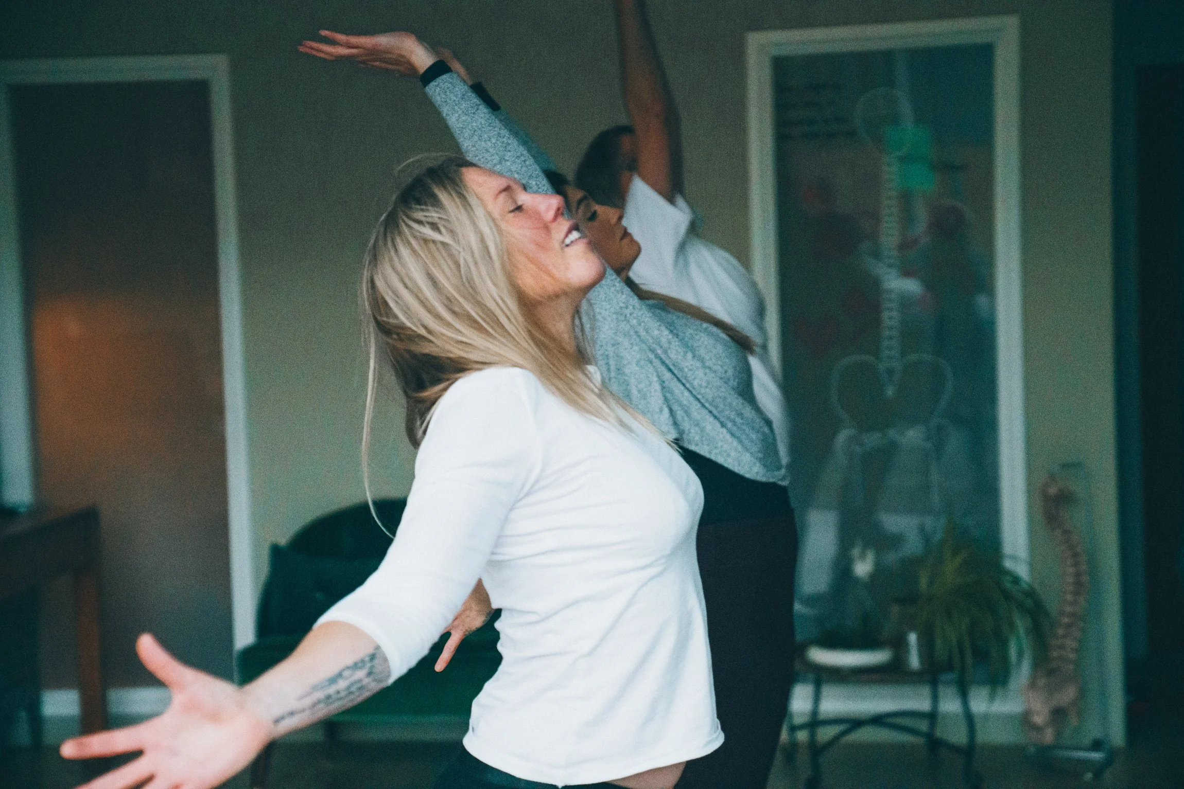 Abi Adams is posing with her arms outstretched behind her, and her eyes are closed towards the ceiling. Two other people are visible in the background with their arms towards the ceiling.