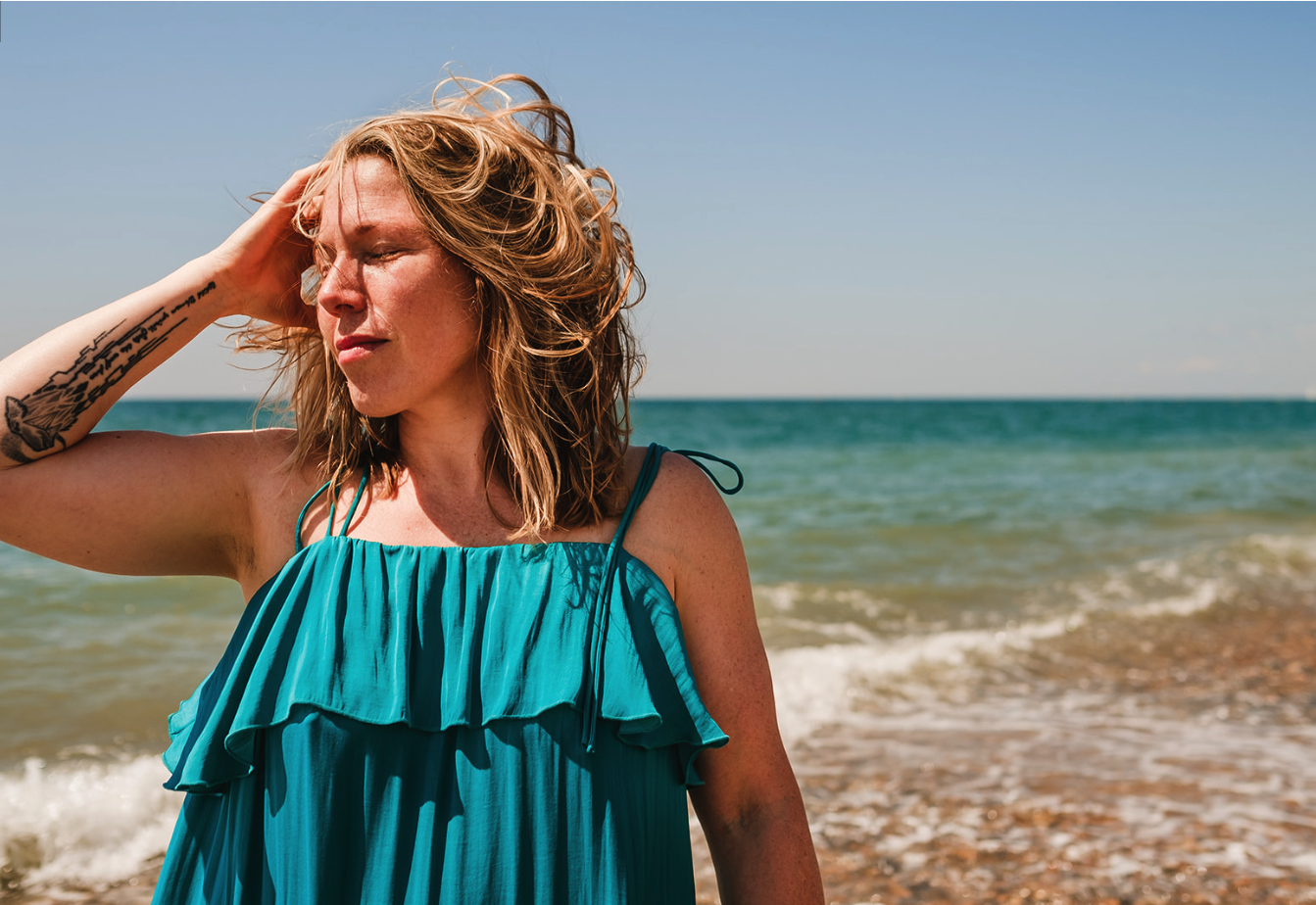 Abi Adams is standing in front of the sea at a beach, she is wearing a blue dress and is running one hand through her hair.