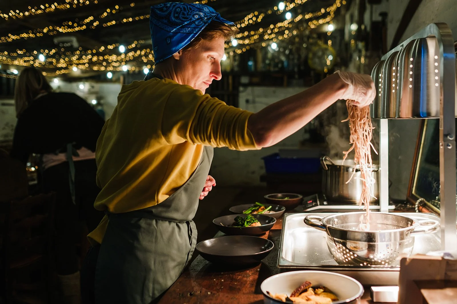 A woman wearing a blue headscarf is standing by a table that has plates on it. She is touching food that is just off camear. Behind her are fairy lights and seats.