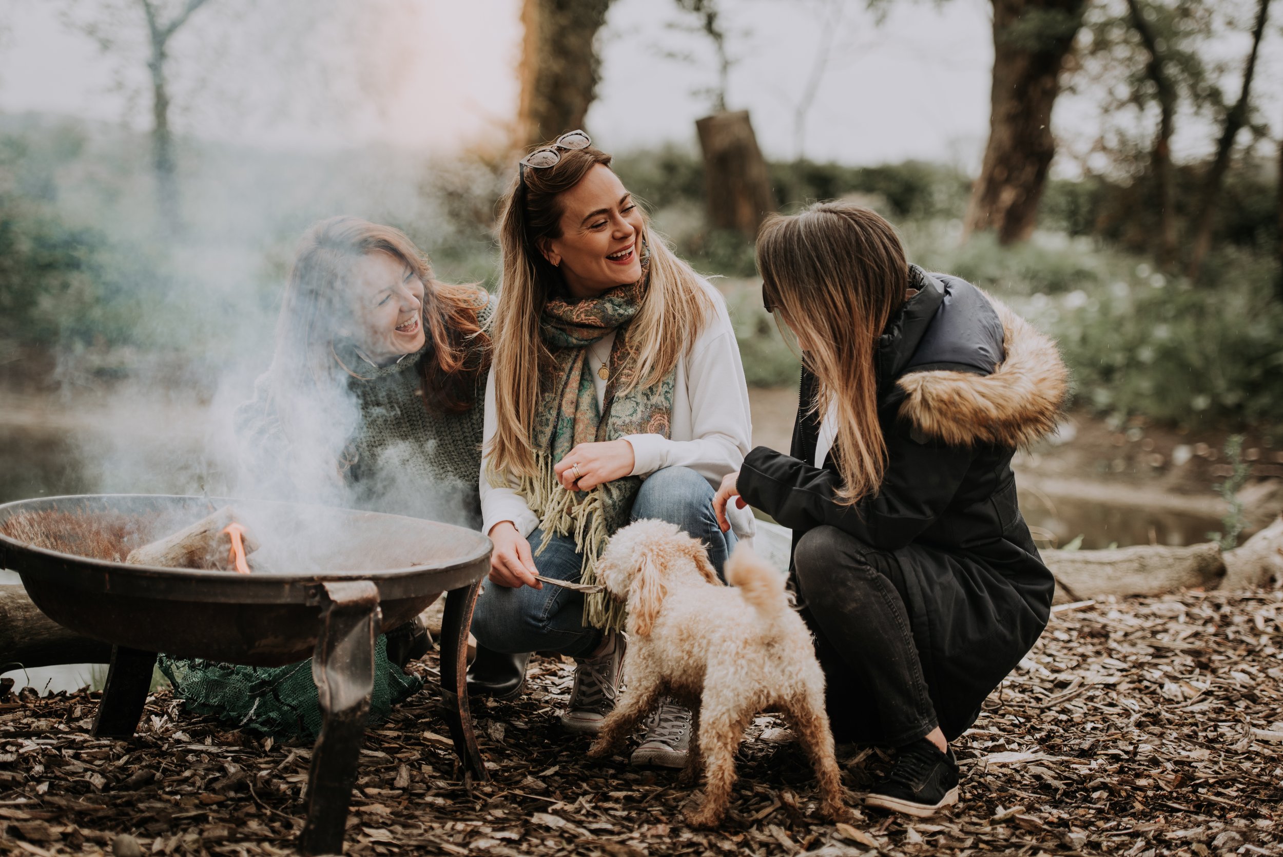A group of women and a dog are sat by a firepit, all the women are laughing and looking towards each other.