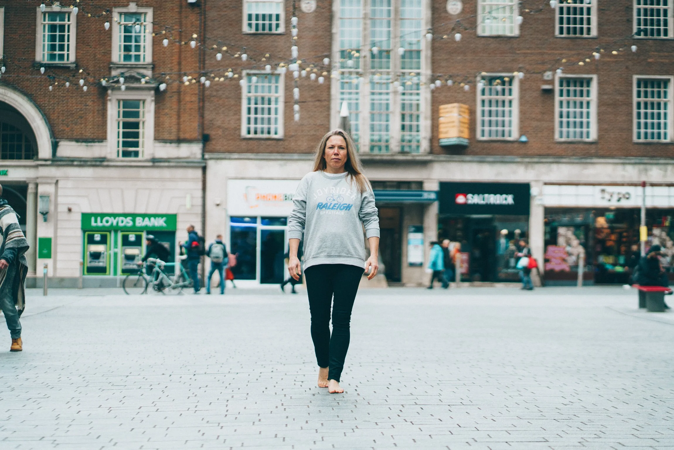 Abi Adams is outside on a pavement walking towards the camera, she is standing in front of a street of shops and restaurants.