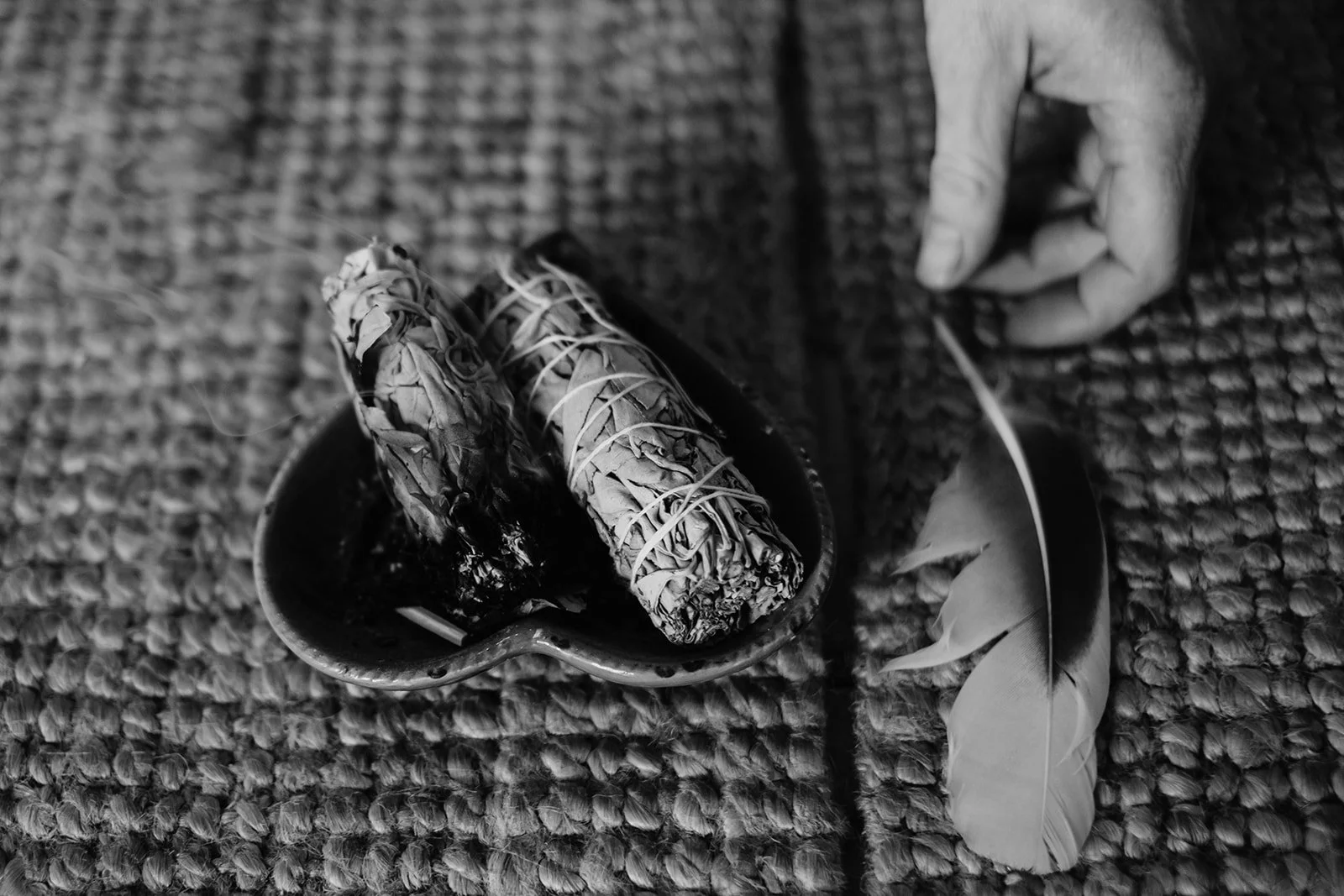 A black and white image of a heart shaped dish with two bundles of sage in it. To the right of the dish is a hand holding a feather.
