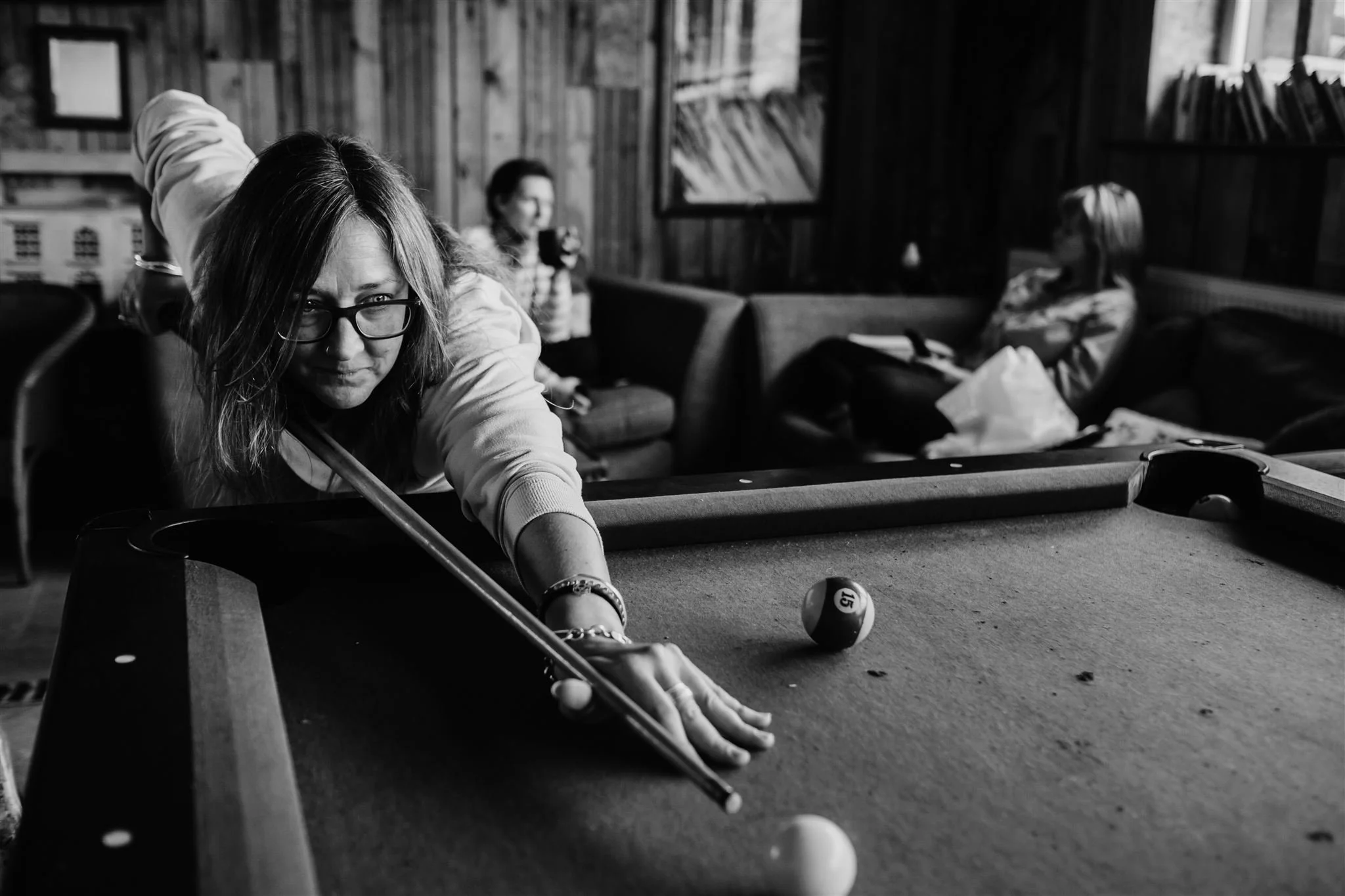 A black and white image of a woman playing pool on a pool table, with two other people visible in the background sitting on chairs.