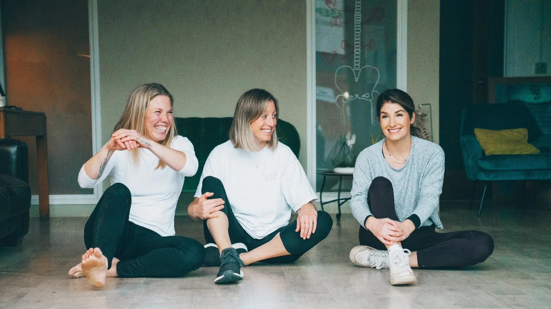 Abi Adams sitting with two other woman on a floor. All of the women are smiling and looking away from the camera.